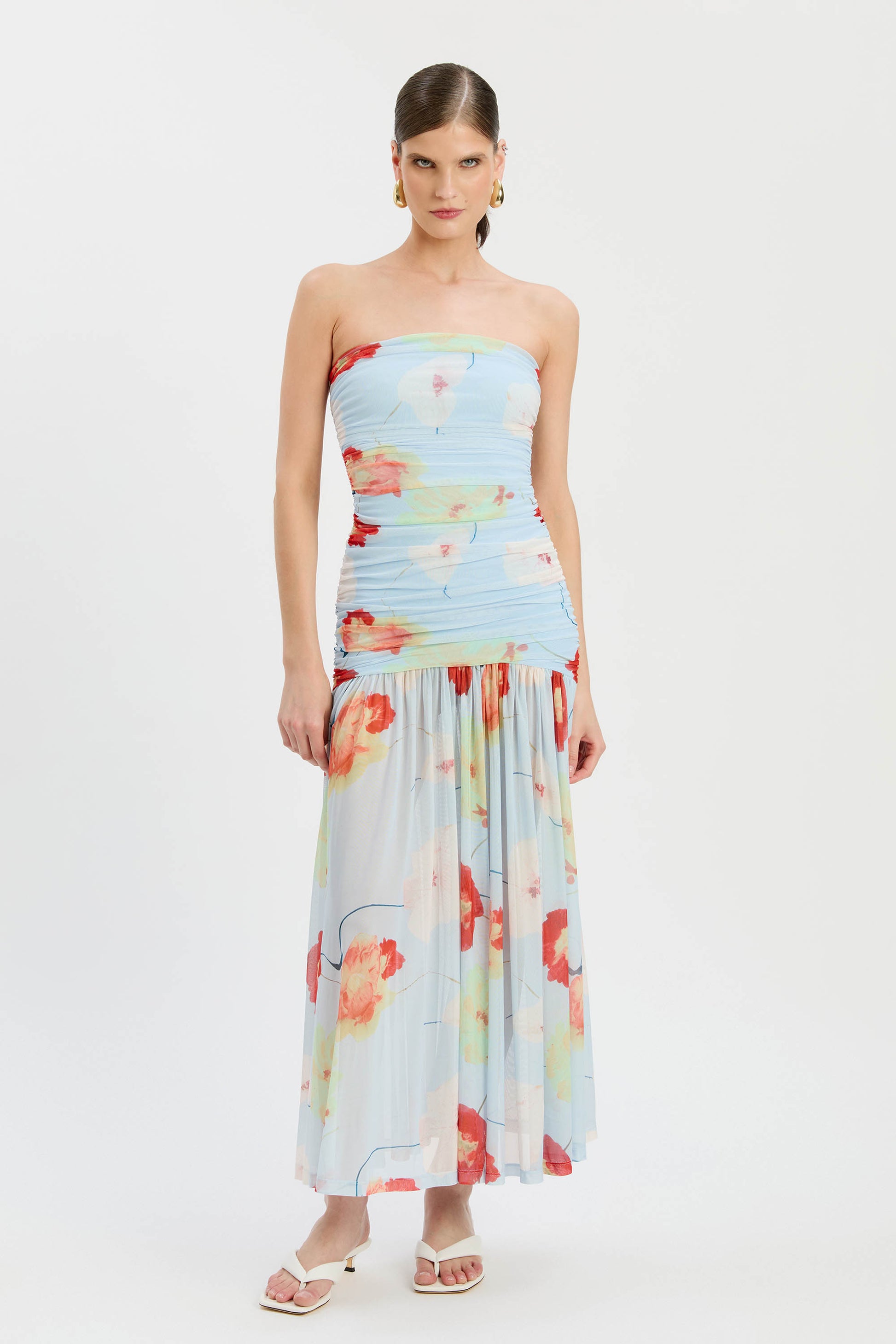 A woman wears the Bardot Solana Printed Mesh Maxi Dress, featuring a strapless ruched bodice and flowy ankle-length mesh skirt. Her hair is slicked back, and she accessorizes with gold earrings and white sandals.