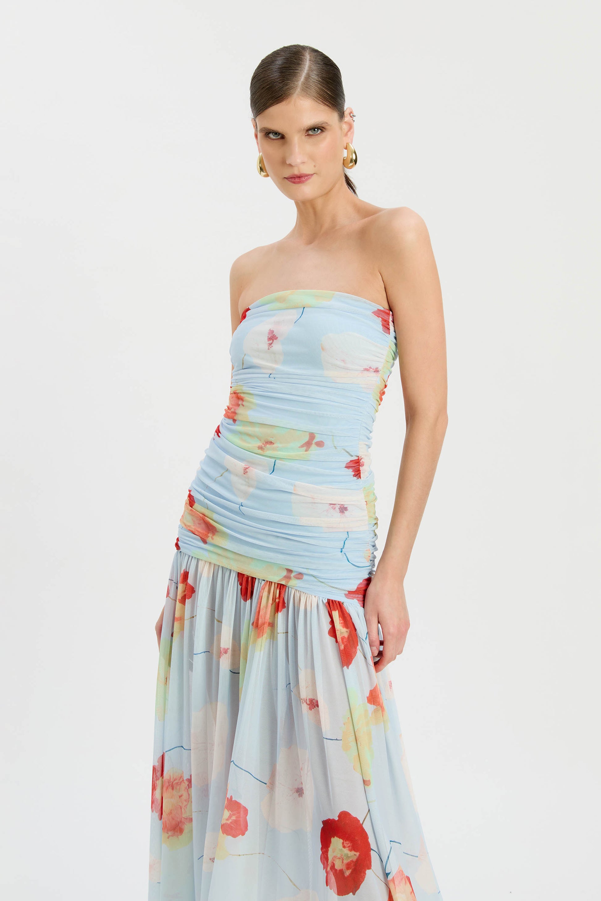 A woman wears the Bardot Solana Printed Mesh Maxi Dress—slim fit, strapless, light blue with red, orange, and yellow florals—with gold hoop earrings, posing against a plain white background.