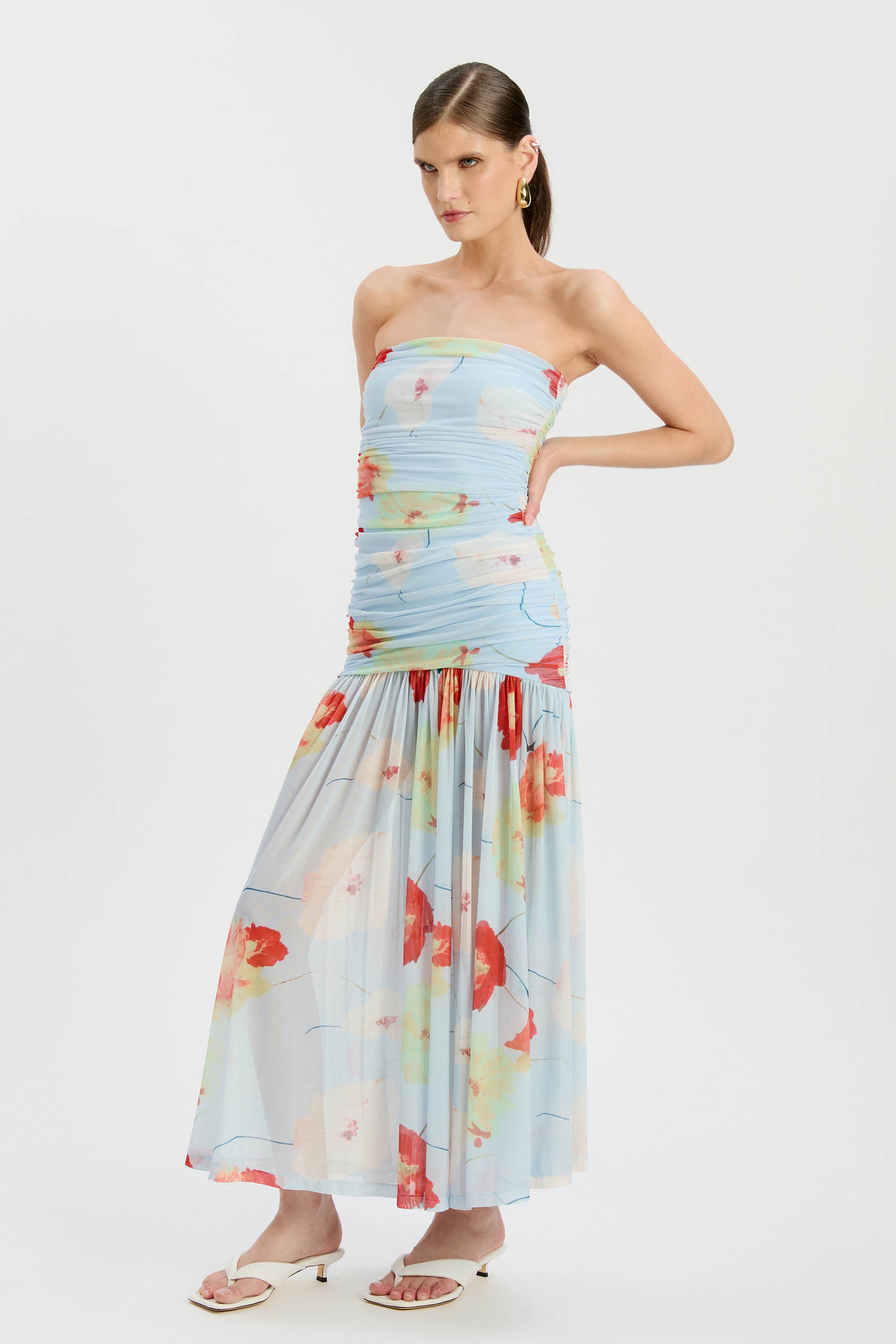 A woman poses against a plain white background wearing the Bardot Solana Printed Mesh Maxi Dress, featuring a strapless ruched bodice, light blue mesh with red-orange floral prints, slim fit top, breezy skirt, white heeled sandals, and gold earrings.