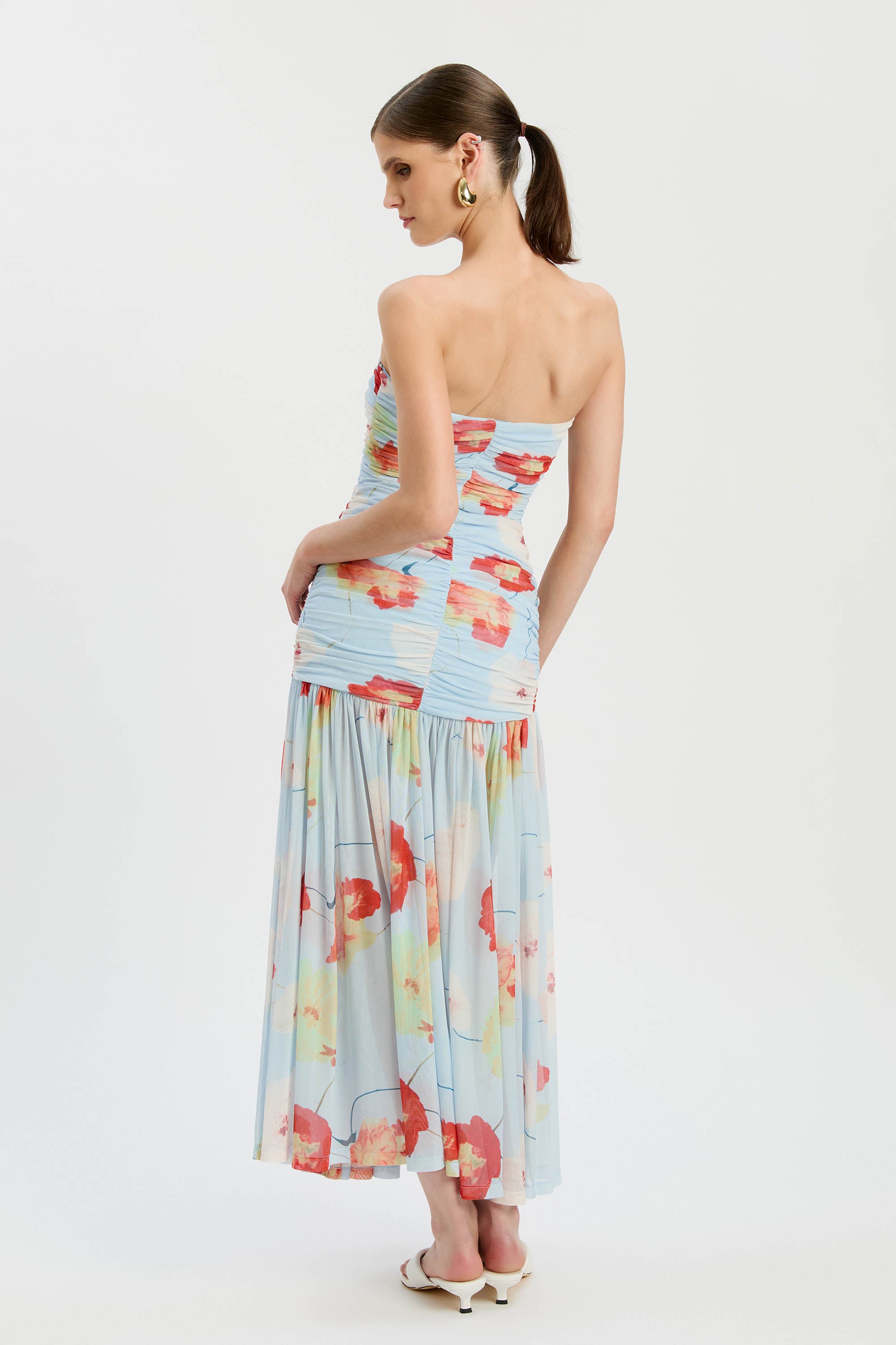 A woman with brown hair in a low ponytail wears the Bardot Solana Printed Mesh Maxi Dress, featuring a strapless slim-fit bodice and flared skirt in blue mesh with red and orange prints. She pairs it with earrings and white sandals against a plain background.
