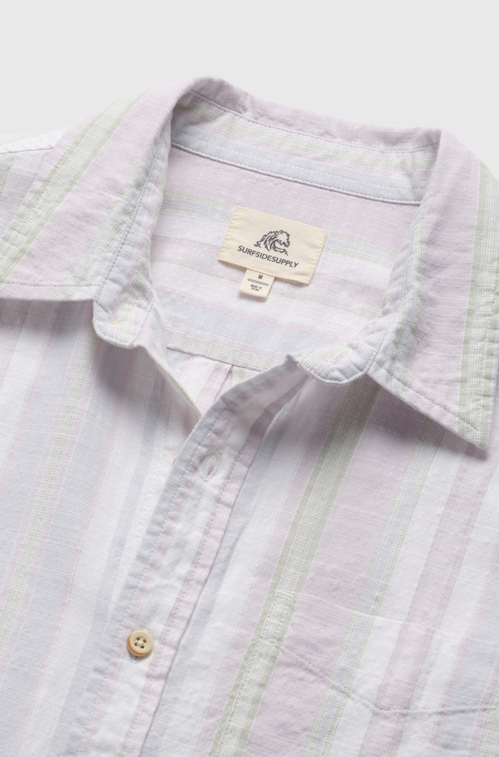 A close-up of the Joey 2 Tone Stripe Slub Shortsleeve shirt by Surfside Supply Co., showing pale green and lavender vertical stripes, lightweight cotton fabric, a collar, white buttons, and a visible brand label inside the collar.