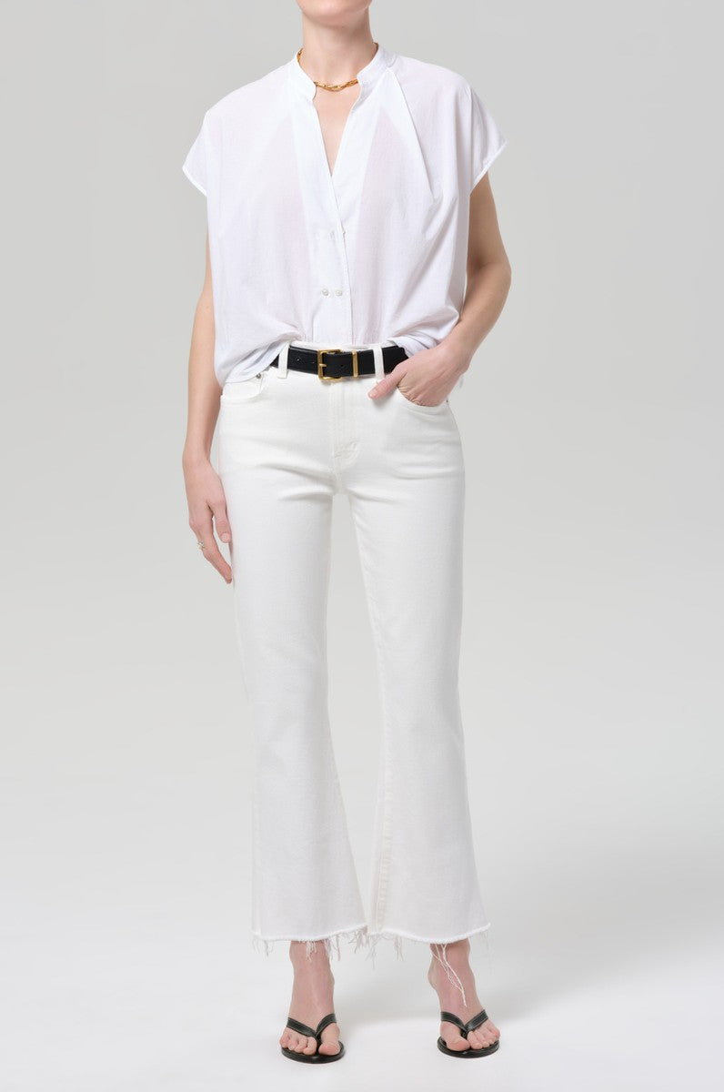 A person stands against a light gray background wearing a short-sleeve white button-up shirt, the Citizens of Humanity Goldie Kick Flare 27 Raw Hem jeans, a black belt, black heeled sandals, and a gold necklace.