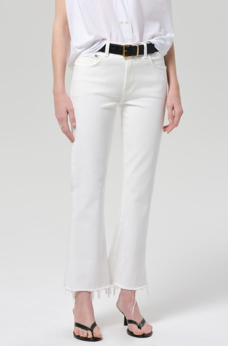 A person in Citizens of Humanity Goldie Kick Flare 27 Raw Hem white jeans, paired with a black belt, tucked-in white shirt, and black heeled sandals stands against a plain light background.