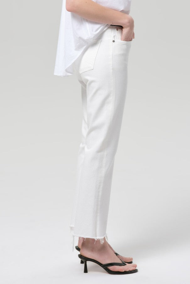 A person stands in profile against a plain light gray background wearing Citizens of Humanity Goldie Kick Flare 27 Raw Hem jeans, a white top, and black heeled sandals.