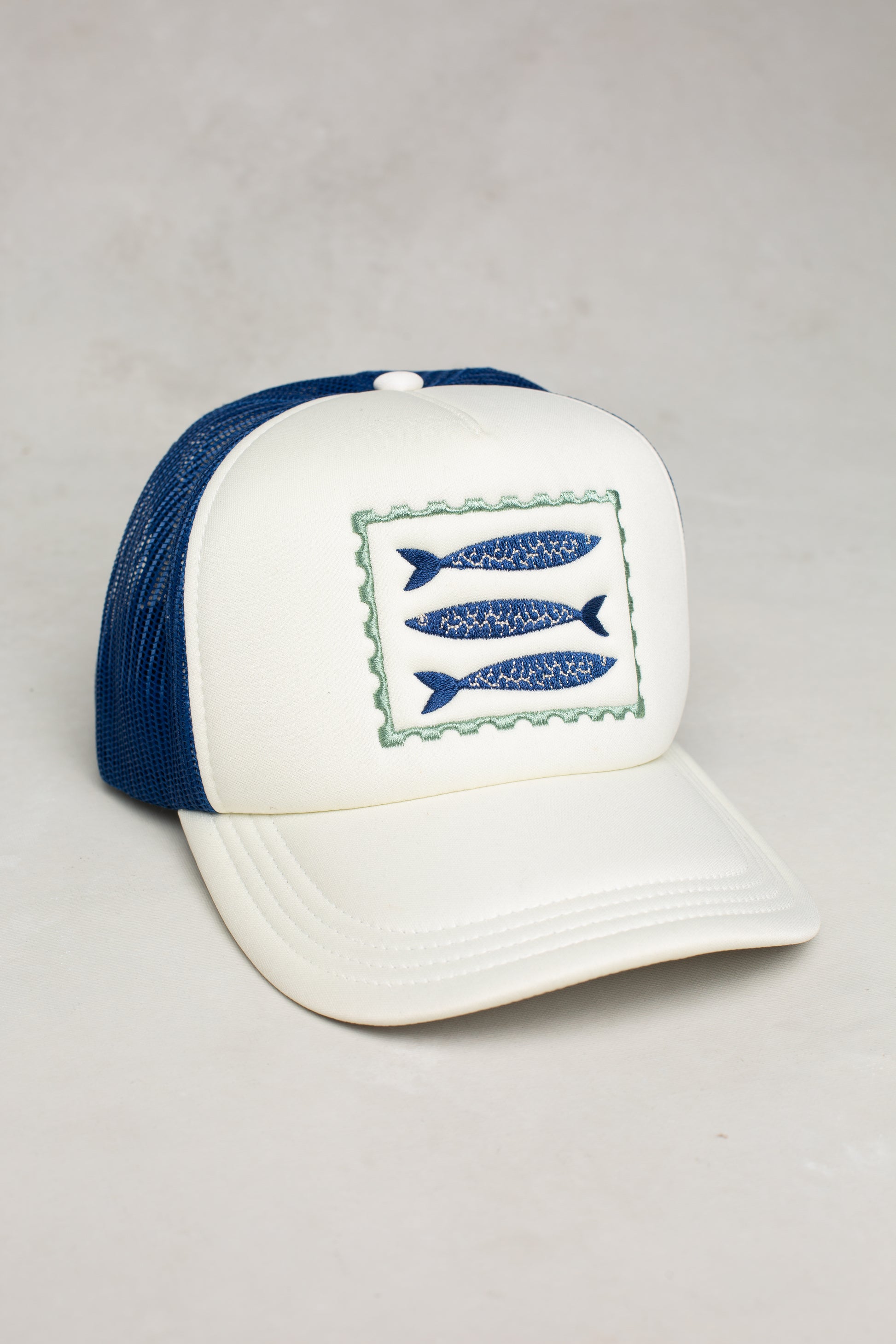 The Sardines Coastal Trucker Hat by Isla x Tate features a white and navy design with a patch of three blue fish, a breathable mesh back, and an adjustable snapback closure. Shown on a light, neutral background.
