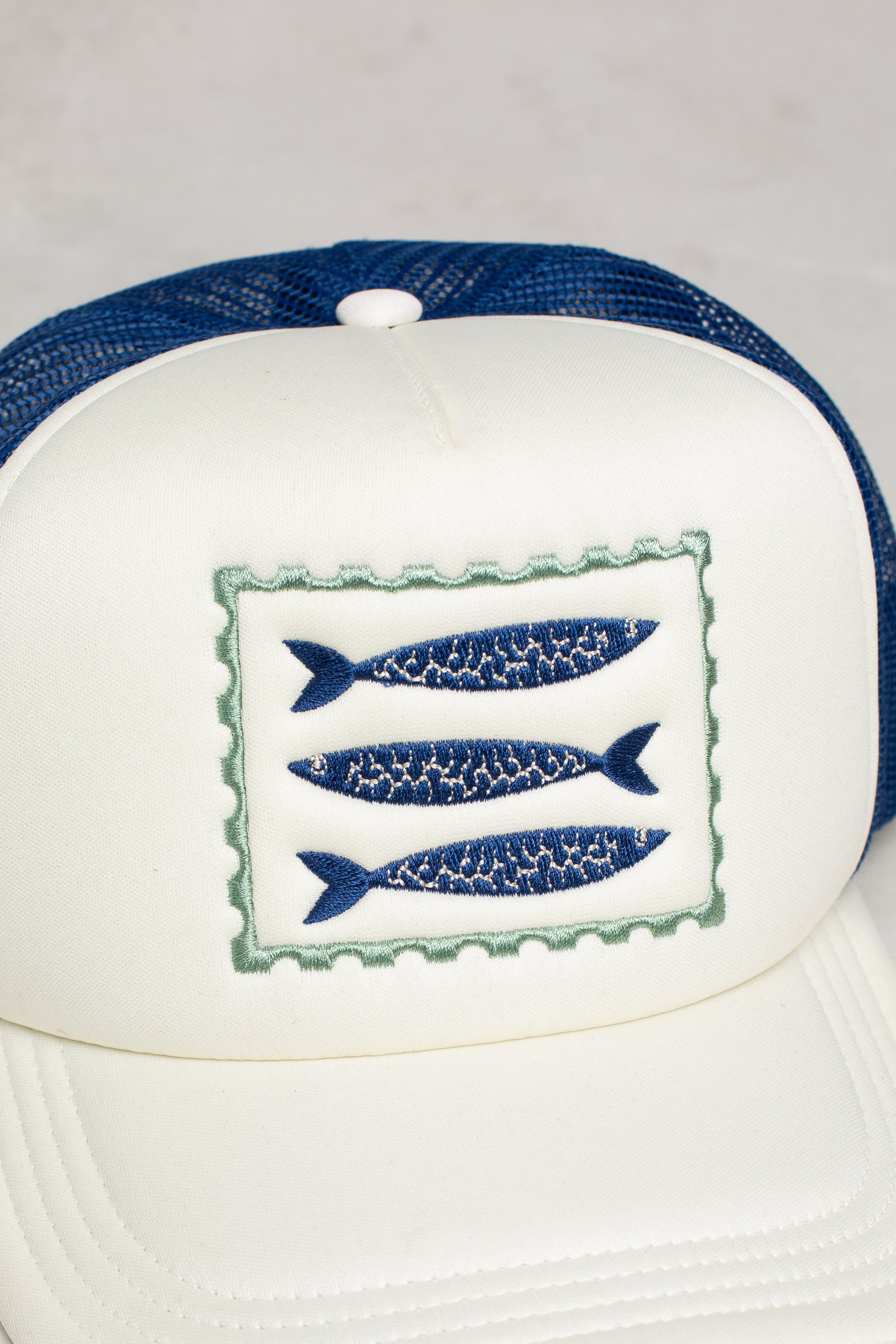The Isla x Tate Sardines Coastal Trucker Hat is a white and blue mesh-back hat with a front patch of three blue fish, offering breathable comfort all day.
