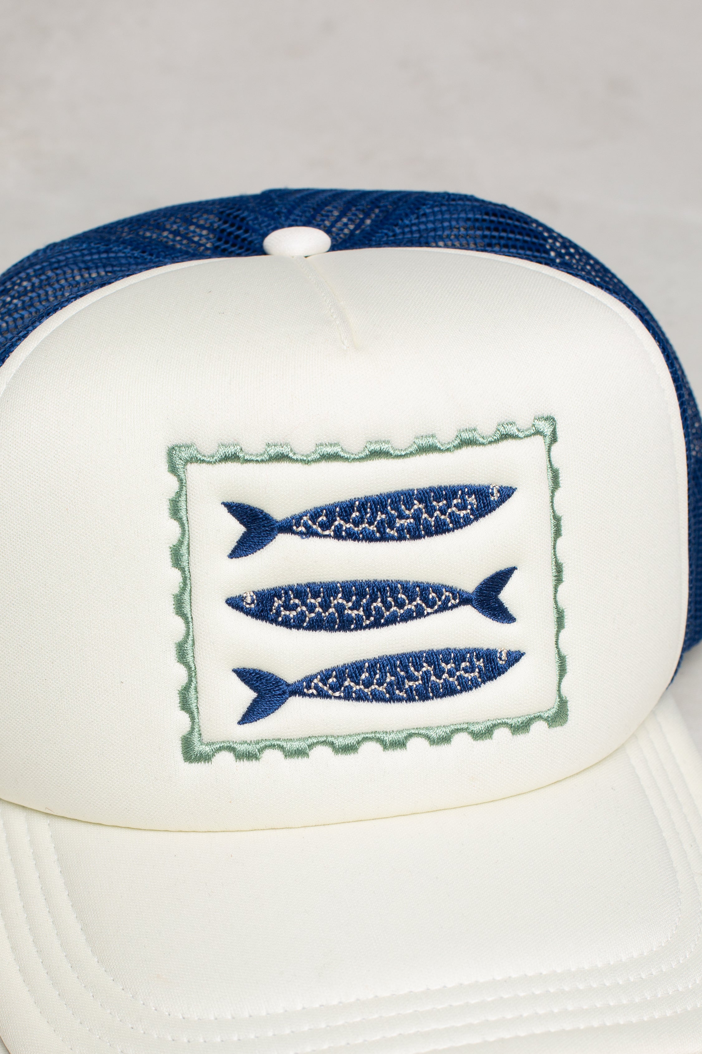 The Isla x Tate Sardines Coastal Trucker Hat is a white and blue mesh-back hat with a front patch of three blue fish, offering breathable comfort all day.