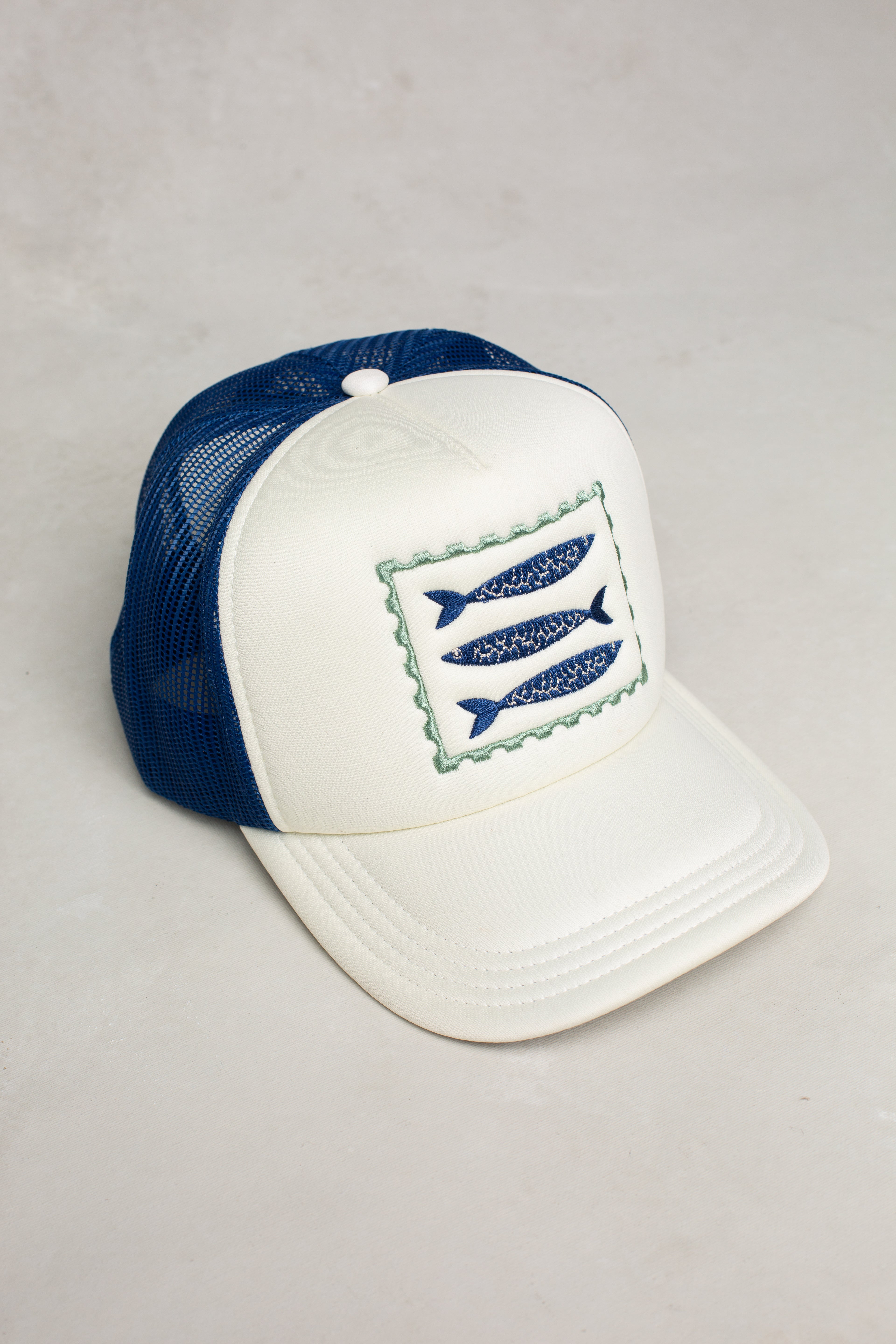 The Isla x Tate Sardines Coastal Trucker Hat is a white and blue mesh-back cap with breathable comfort. It features an embroidered patch of three blue fish framed by a wavy green, postage stamp-inspired border on the front.