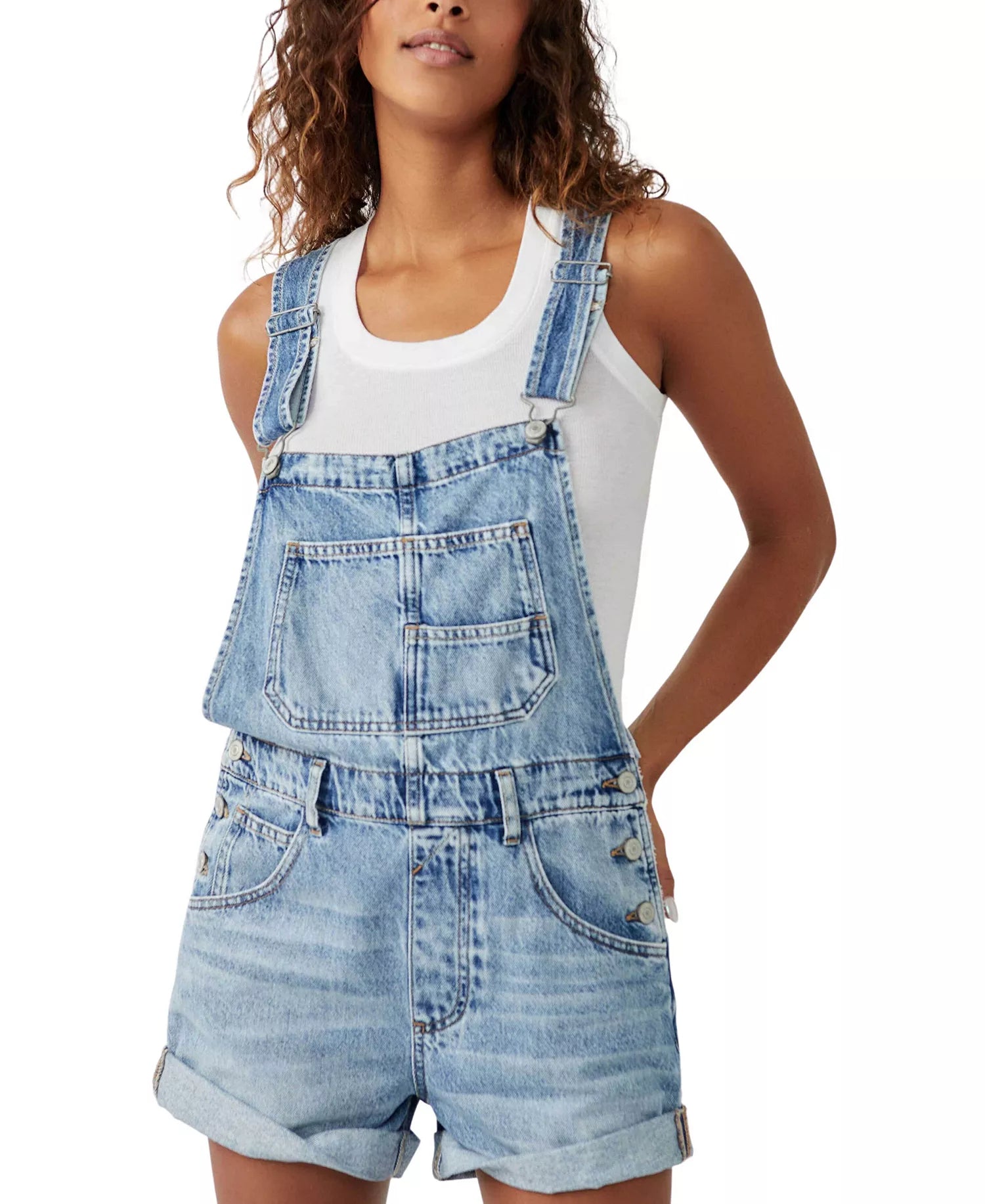 A person models the Free People Ziggy Shortall in light blue denim with adjustable straps over a white sleeveless tank, standing with one hand in a pocket against a plain background.