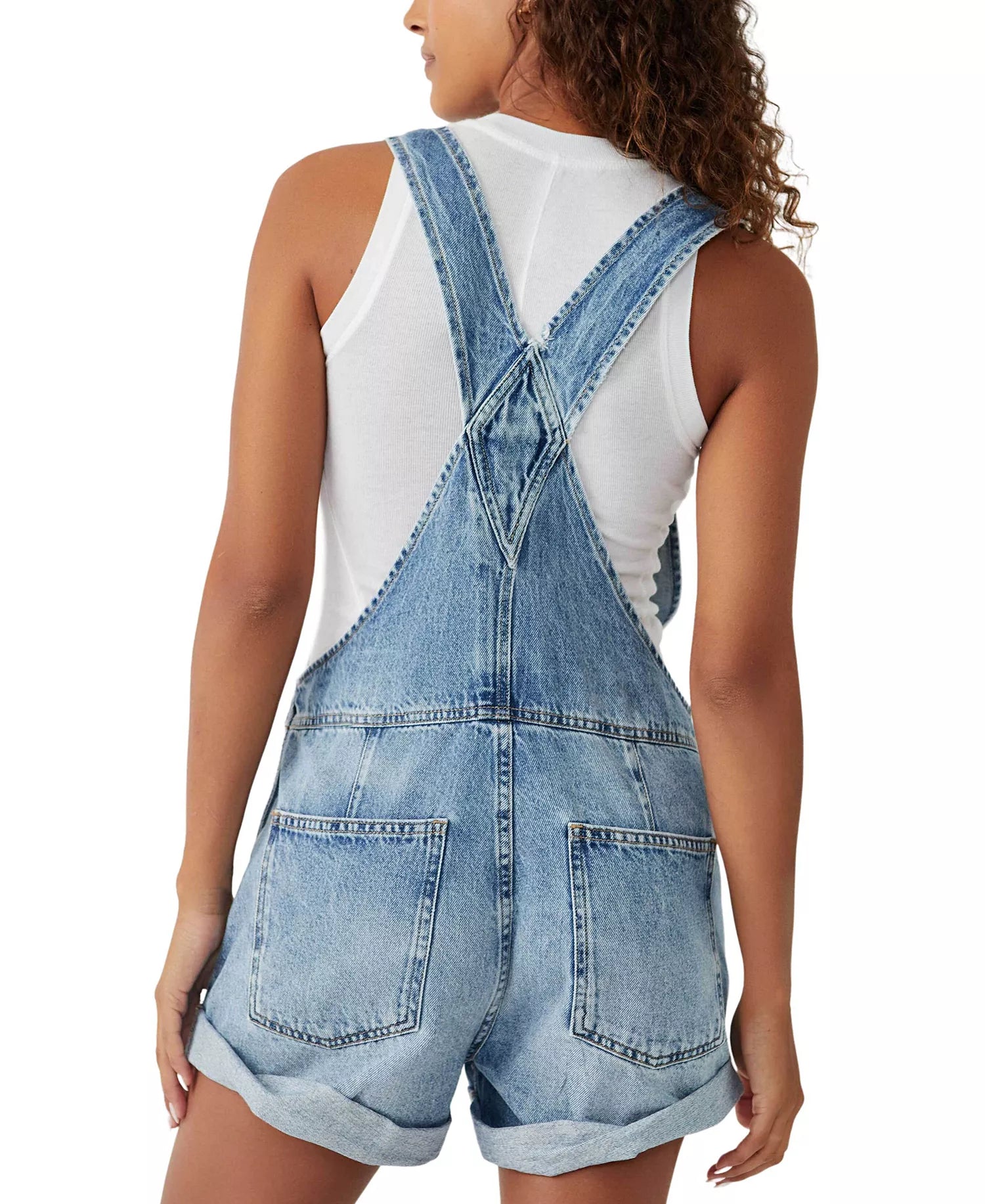 A person with curly hair is shown from the back wearing the Free People Ziggy Shortall, featuring light blue denim, large back pockets, crossed straps, and paired with a white sleeveless top.