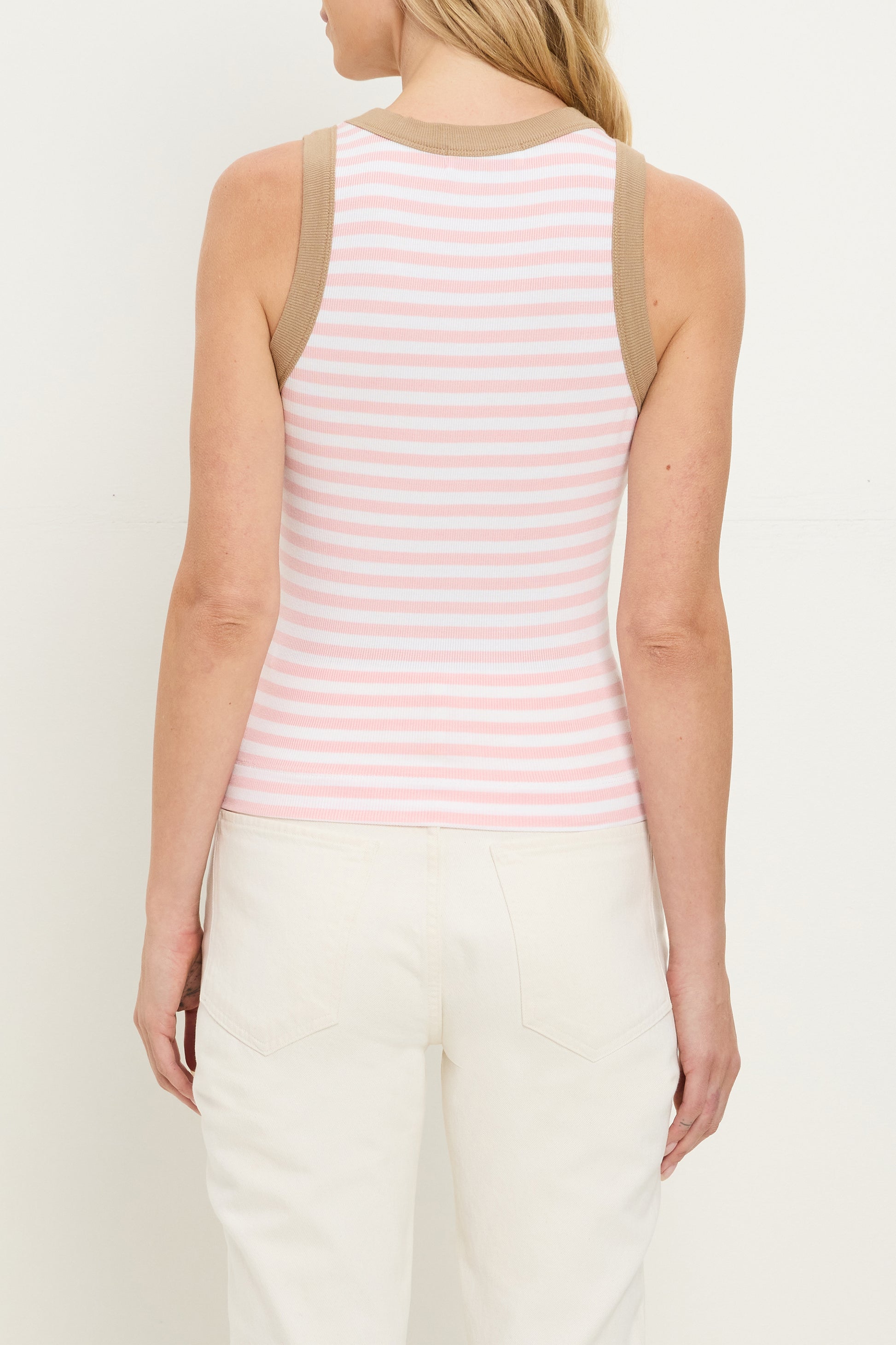 A woman is seen from the back wearing the Michael Stars Gina Ringer Tank, featuring Ultra Rib fabric with pink and white stripes and beige trim, paired with high-waisted white pants.