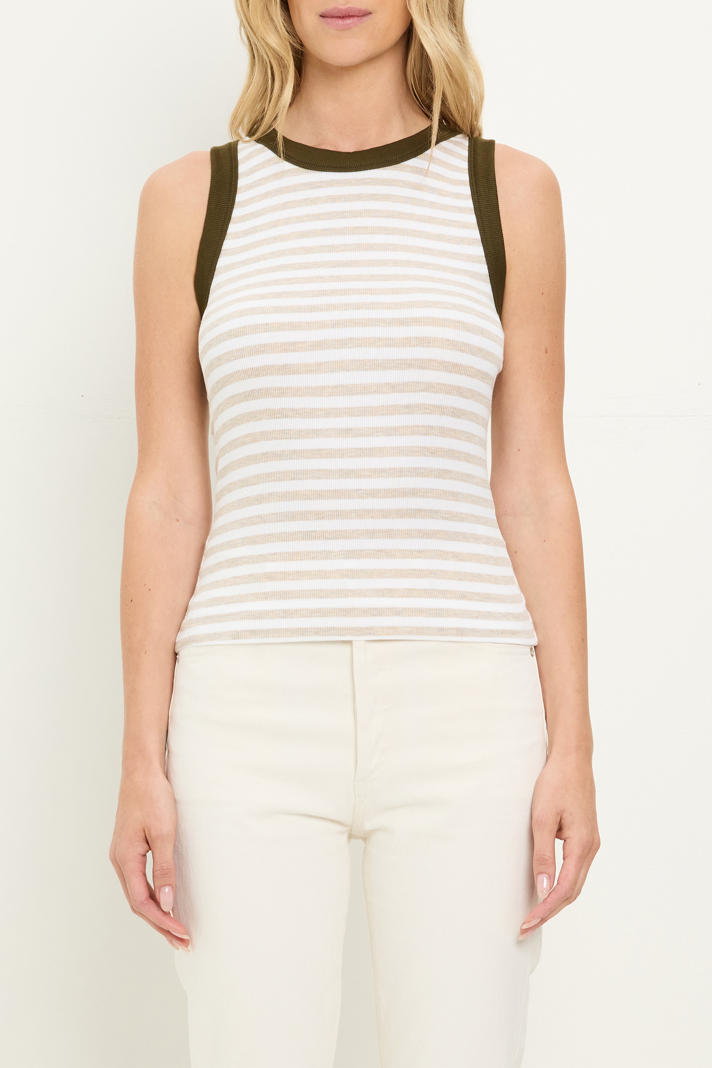 A woman wears the Gina Ringer Tank by Michael Stars, a sleeveless fitted shirt in ultra rib fabric with white and light beige stripes, dark green trim at the neckline, paired with high-waisted off-white pants against a plain light background.