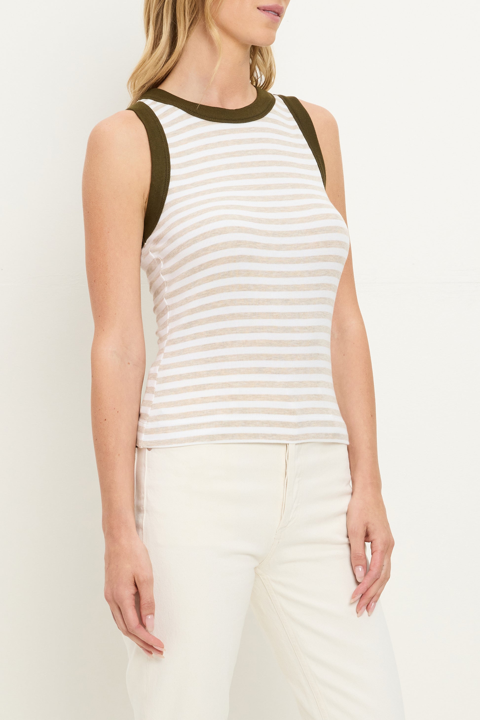 A woman wears the Michael Stars Gina Ringer Tank—a sleeveless white and beige striped top with dark green trim—paired with high-waisted white pants, standing against a plain background.