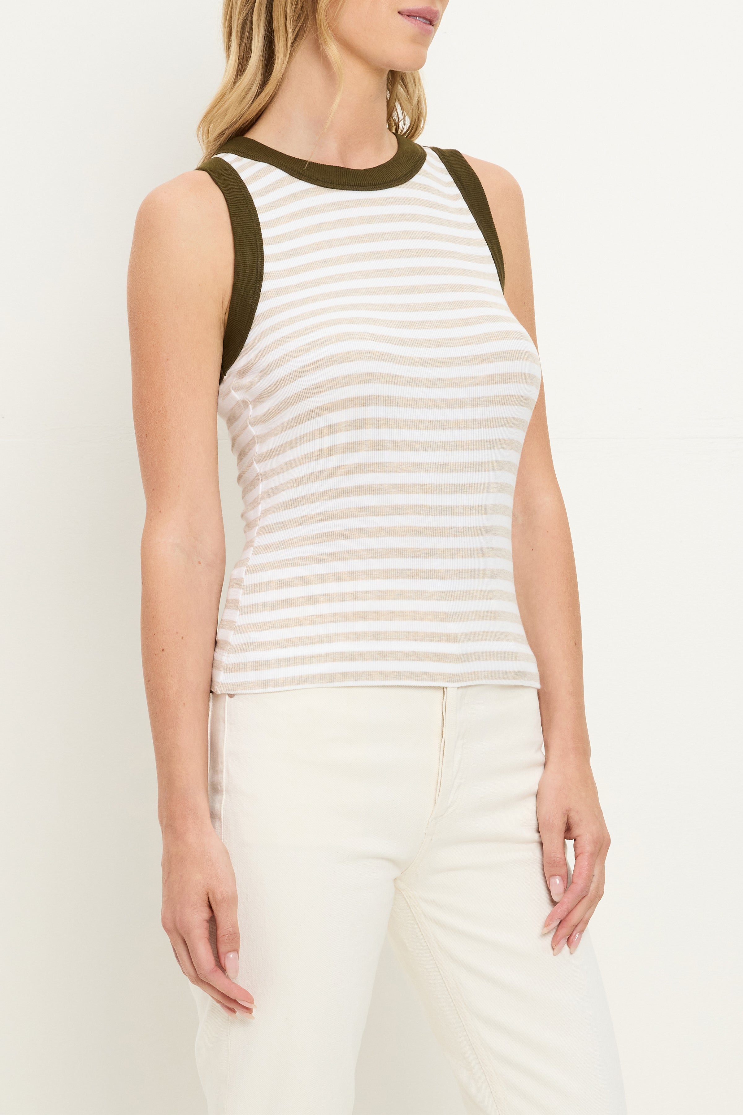 A woman wears the Michael Stars Gina Ringer Tank—a sleeveless white and beige striped top with dark green trim—paired with high-waisted white pants, standing against a plain background.
