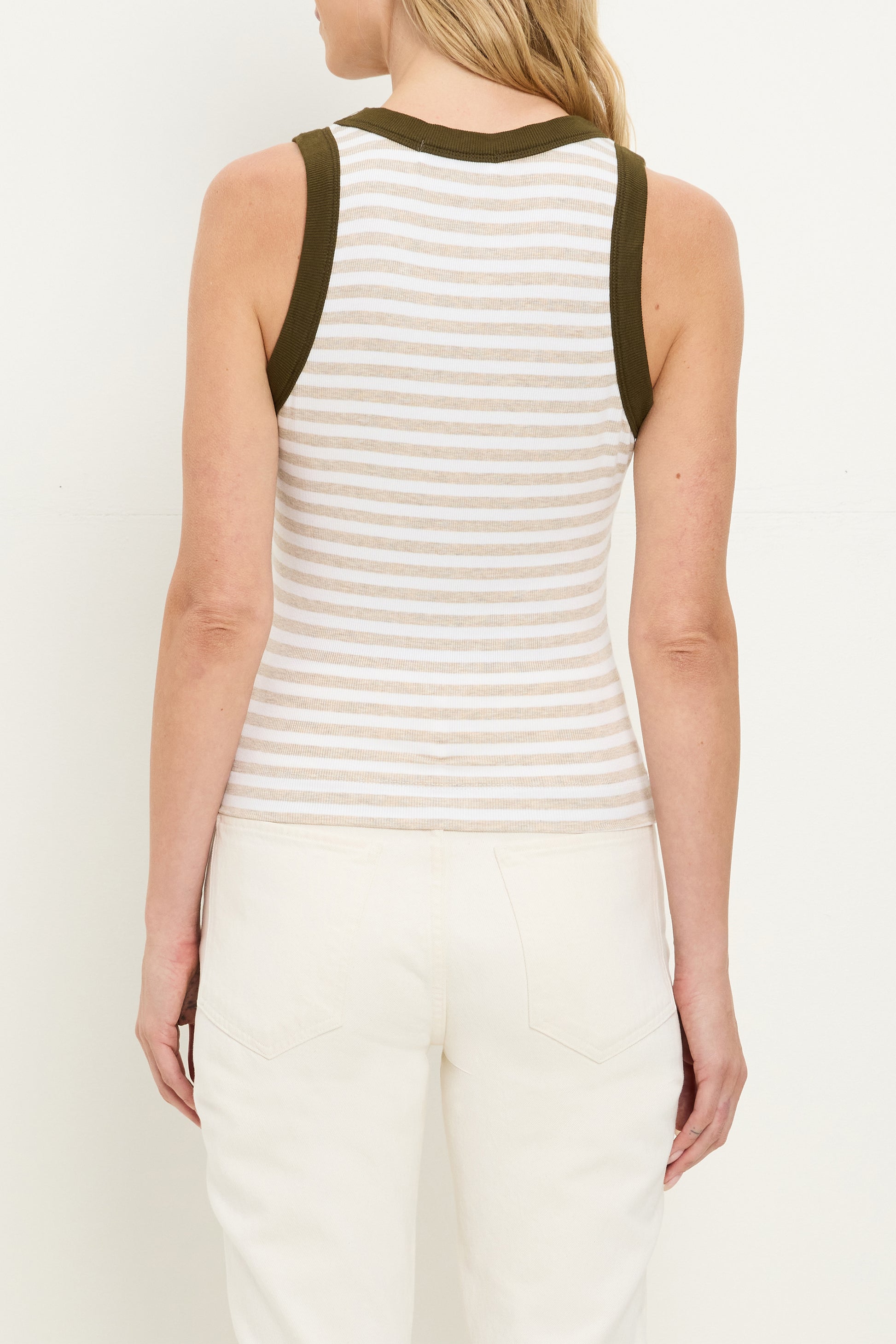A woman with blonde hair and light skin is seen from the back wearing the Michael Stars Gina Ringer Tank, a sleeveless fitted top with white and light gray stripes, dark green trim, and off-white pants.