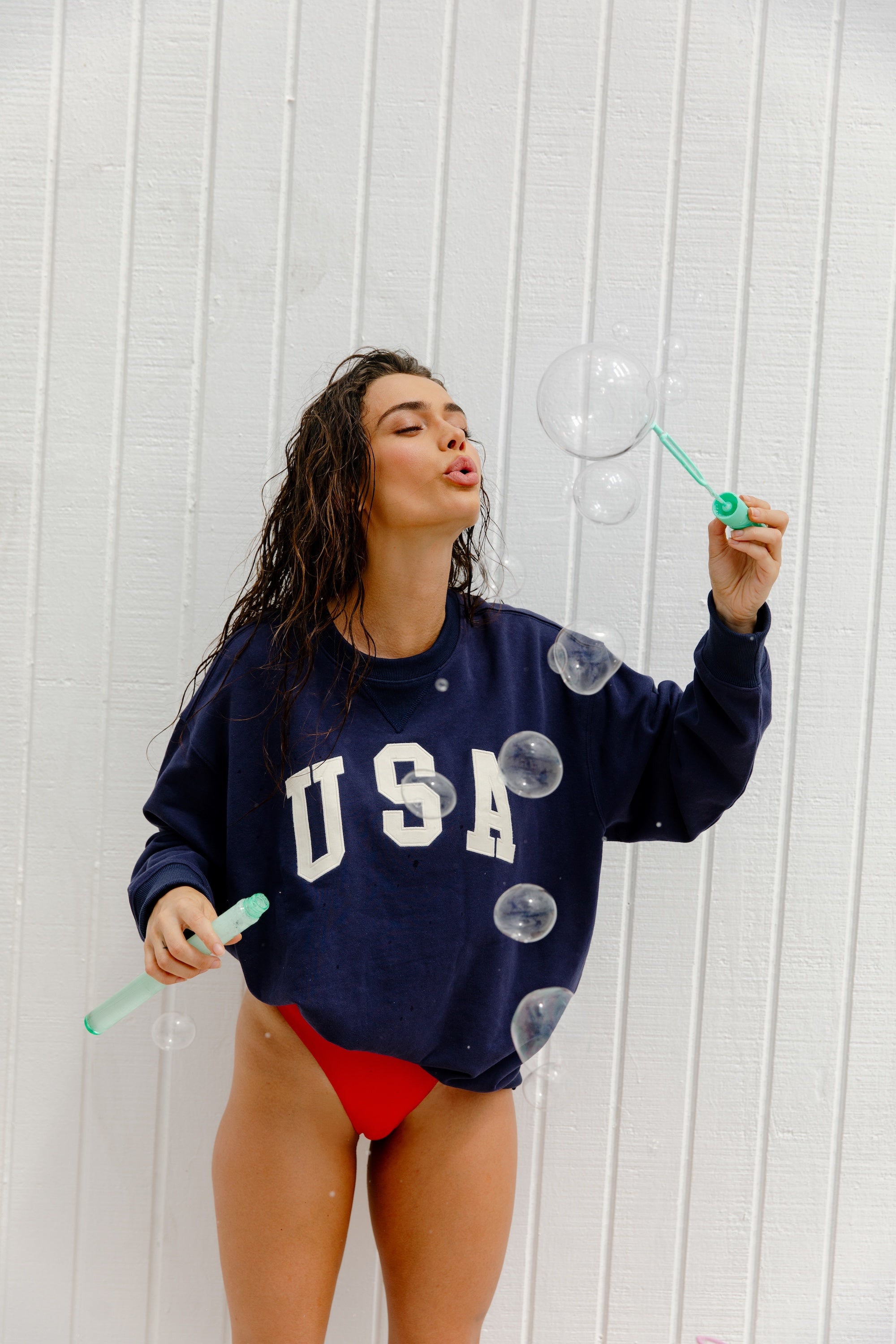 A woman wears the Isla Dolly USA Crew Neck sweatshirt and red bikini bottoms, blowing soap bubbles with wet hair in front of a white wooden wall, holding a bubble wand and tube.