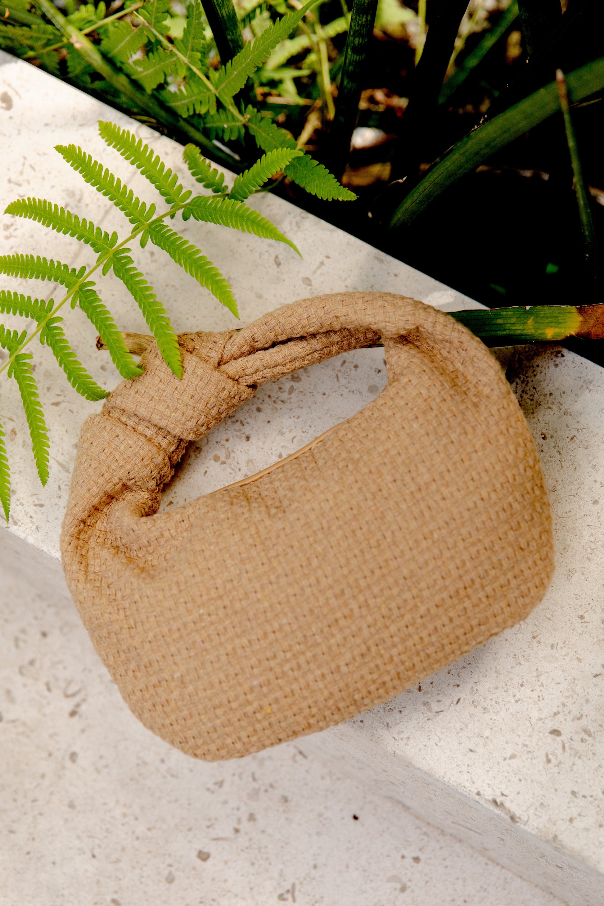 The Tracy Bag in Natural Tweed by Urban Expressions, featuring a knotted top handle and textured weave, rests on a light stone surface beside green fern leaves and tall plants for an earthy, natural vibe.