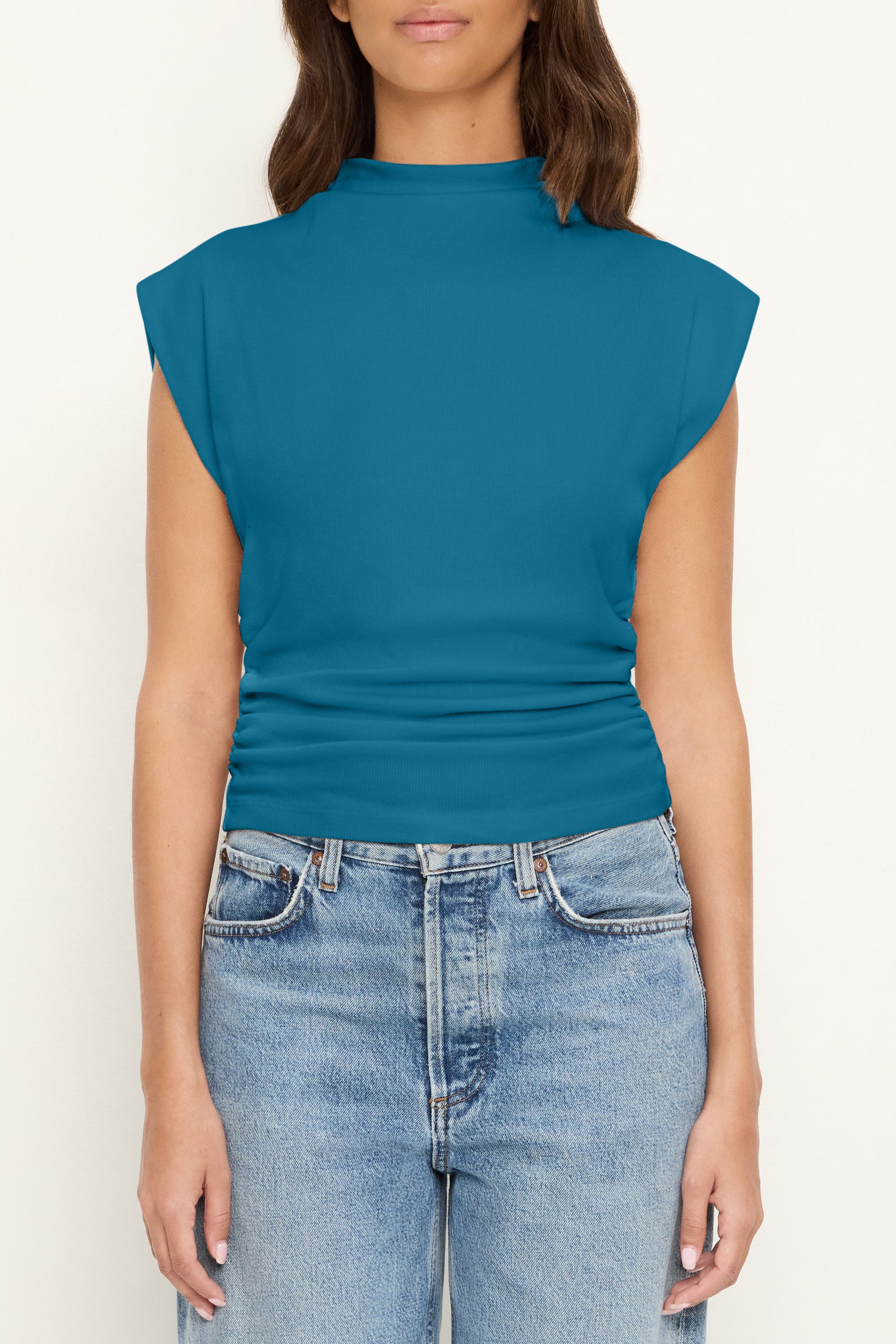A woman stands against a plain light background wearing the Michael Stars Amara Crop Top in Lagoon—a sleeveless, teal mock neck muscle tee—paired with high-waisted blue jeans that accentuate her cinched waist.