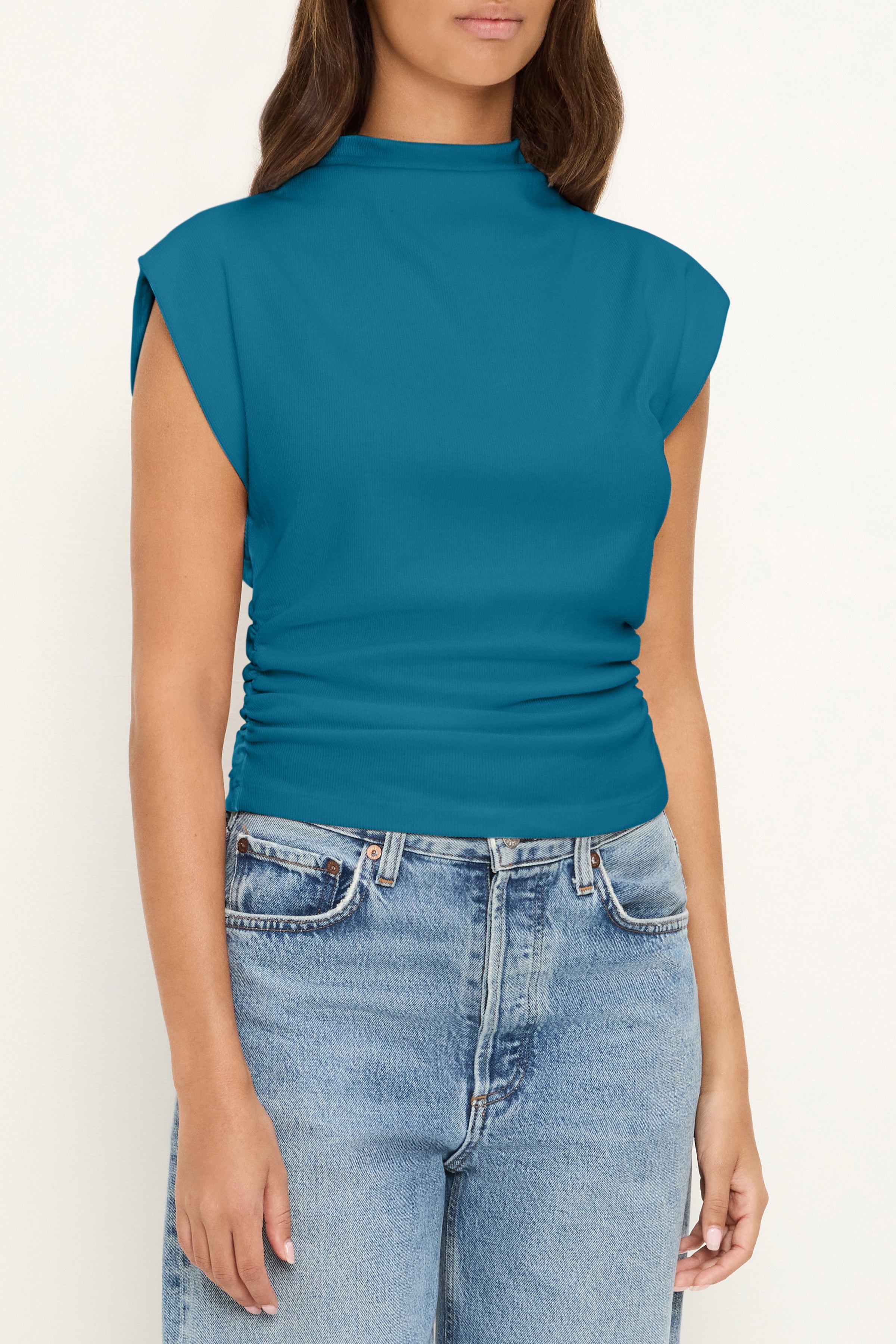 A woman wears the Amara Crop Top In Lagoon by Michael Stars with light blue high-waisted jeans, standing against a plain light background. The cinched waist of her jeans adds definition to the look, and her face is partially visible.
