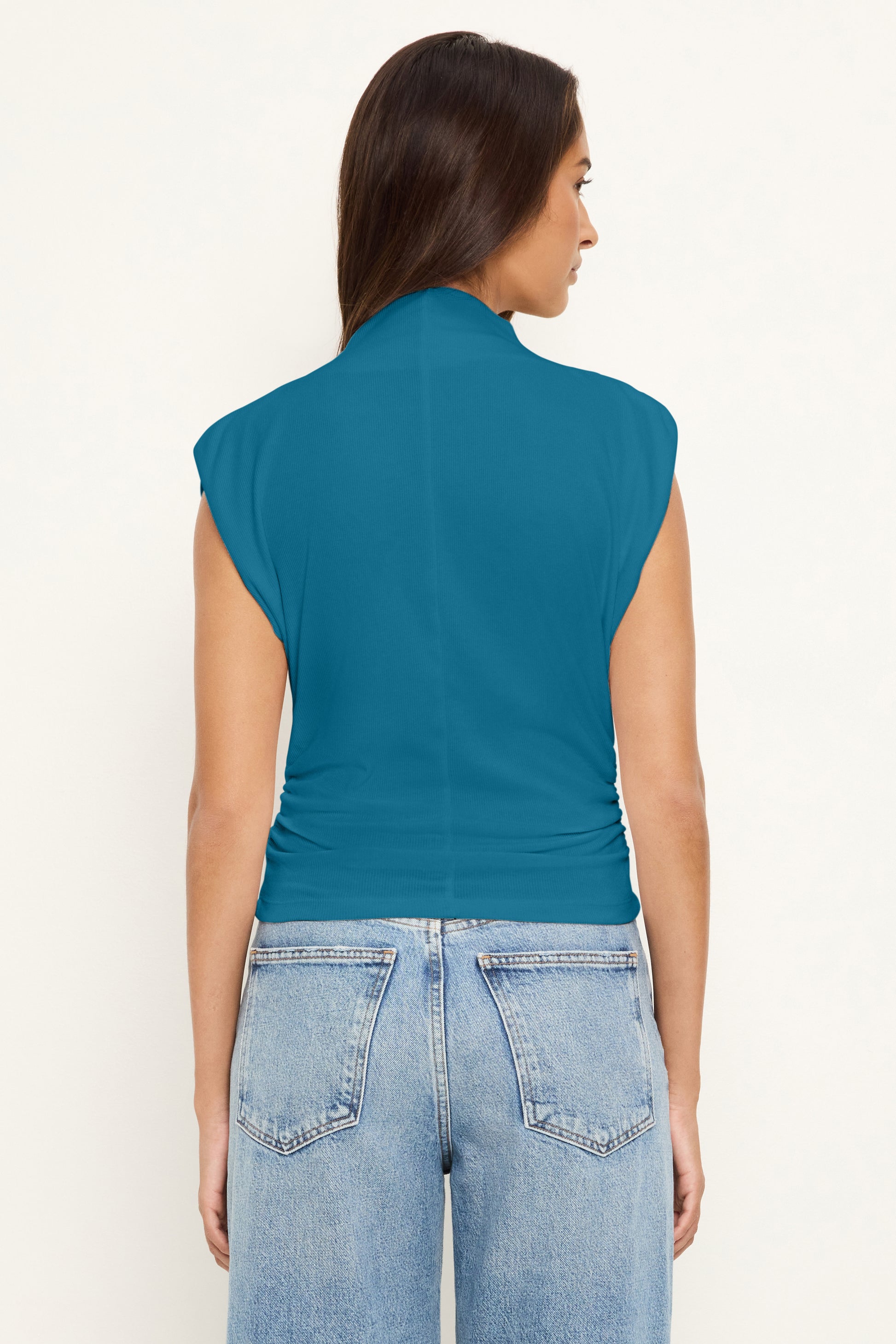 A woman with long brown hair faces away, wearing the Michael Stars Amara Crop Top in Lagoon and light blue high-waisted jeans with a cinched waist against a light background.