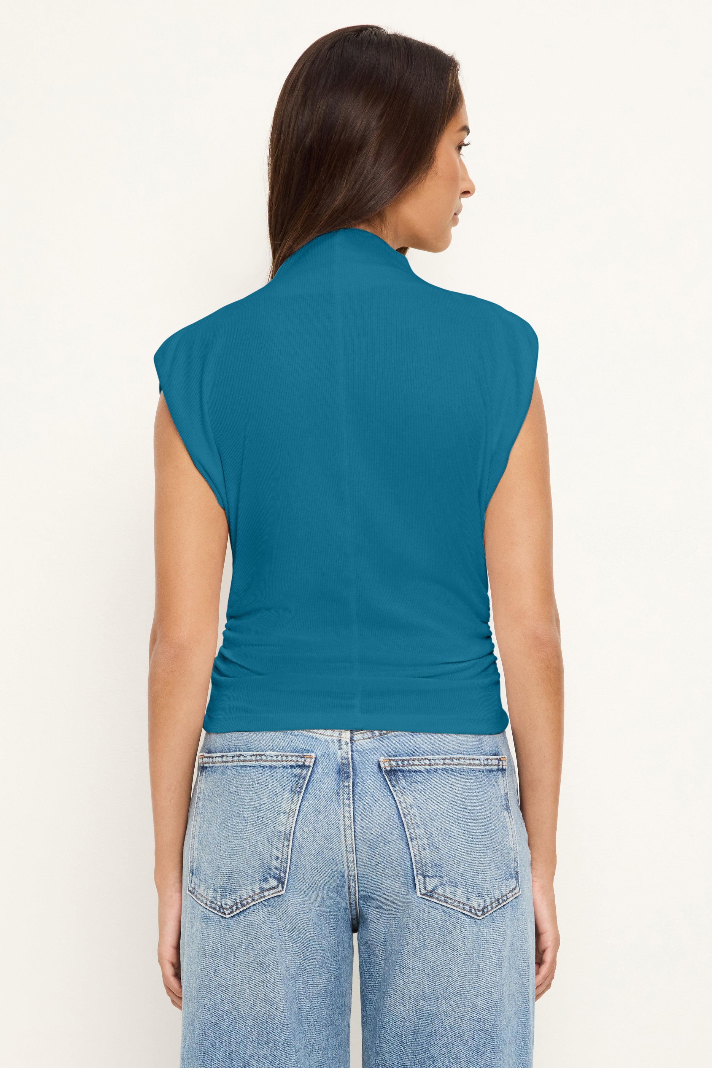 A woman with long brown hair faces away, wearing the Michael Stars Amara Crop Top in Lagoon and light blue high-waisted jeans with a cinched waist against a light background.