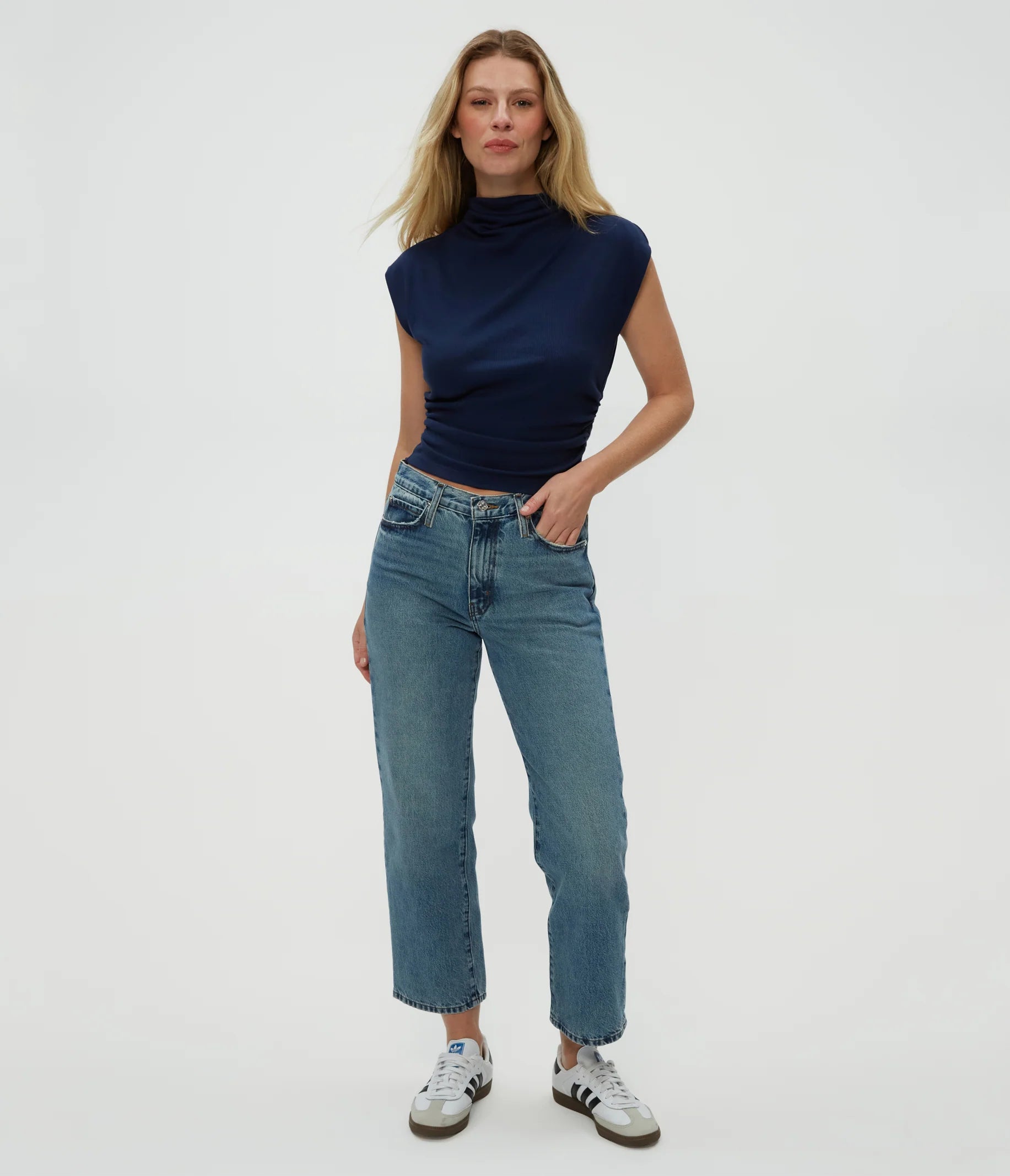 A woman stands confidently in the Michael Stars Amara Crop Top in Nocturnal, paired with blue high-waisted jeans and white sneakers with black accents, against a plain light background.