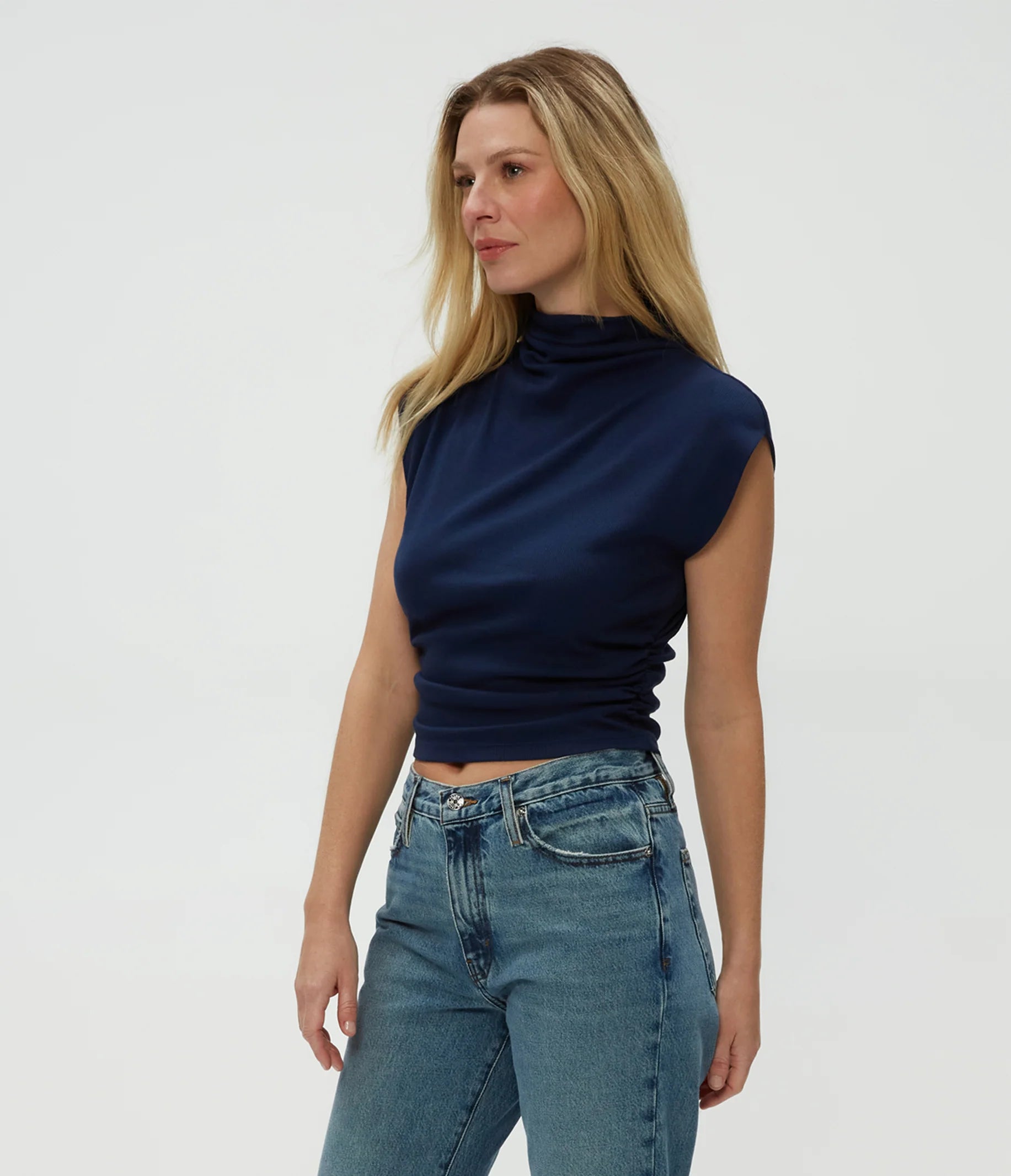 A woman with long blonde hair wears the Michael Stars Amara Crop Top in Nocturnal and high-waisted blue jeans, standing against a plain light background.