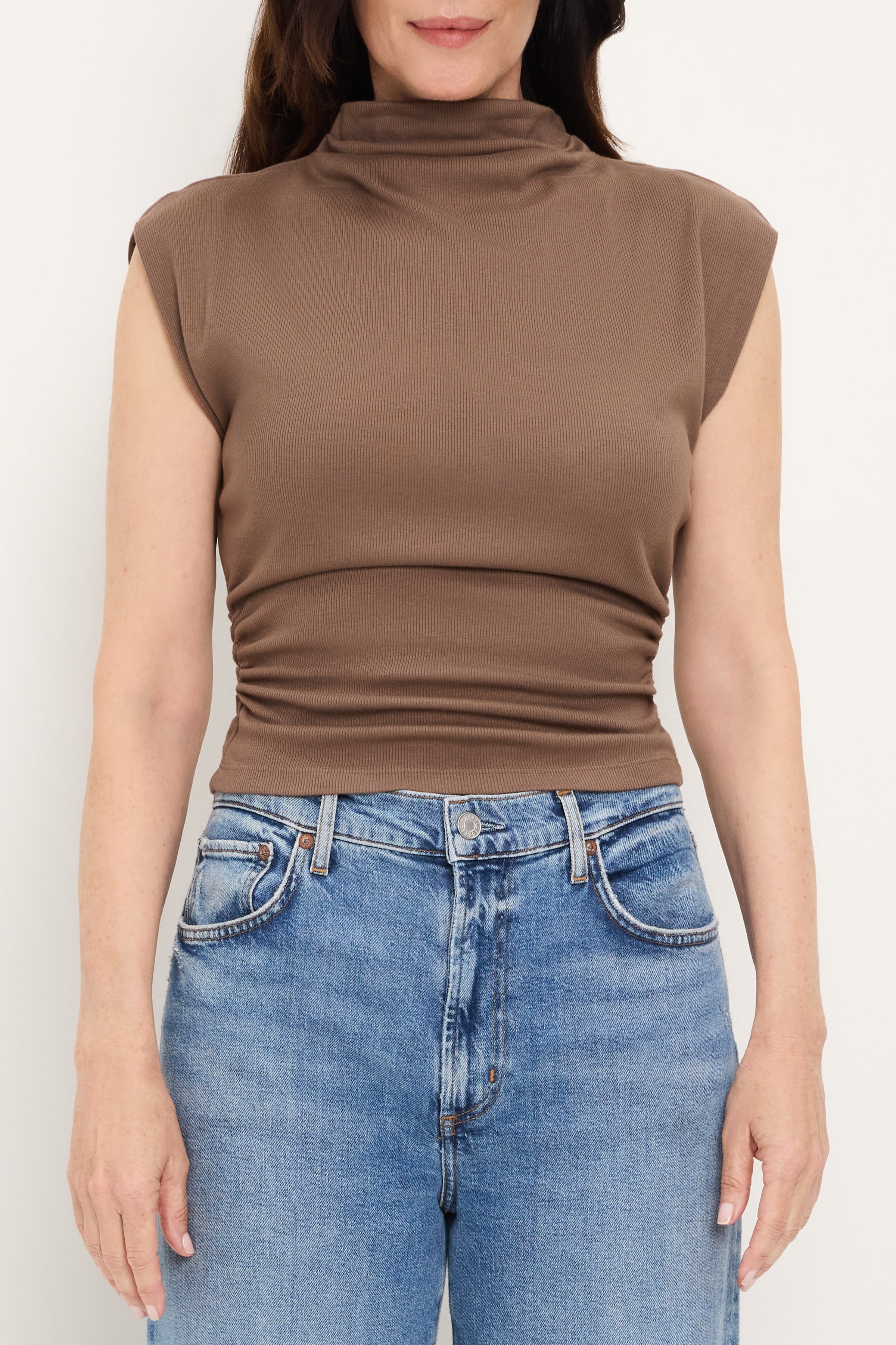 A woman stands against a plain white background wearing the Michael Stars Amara Crop Top in QUICKSAND, a sleeveless mock neck muscle tee made from a soft cotton-modal-spandex blend, paired with blue jeans. Her arms rest relaxed at her sides, and her head is partially out of frame.