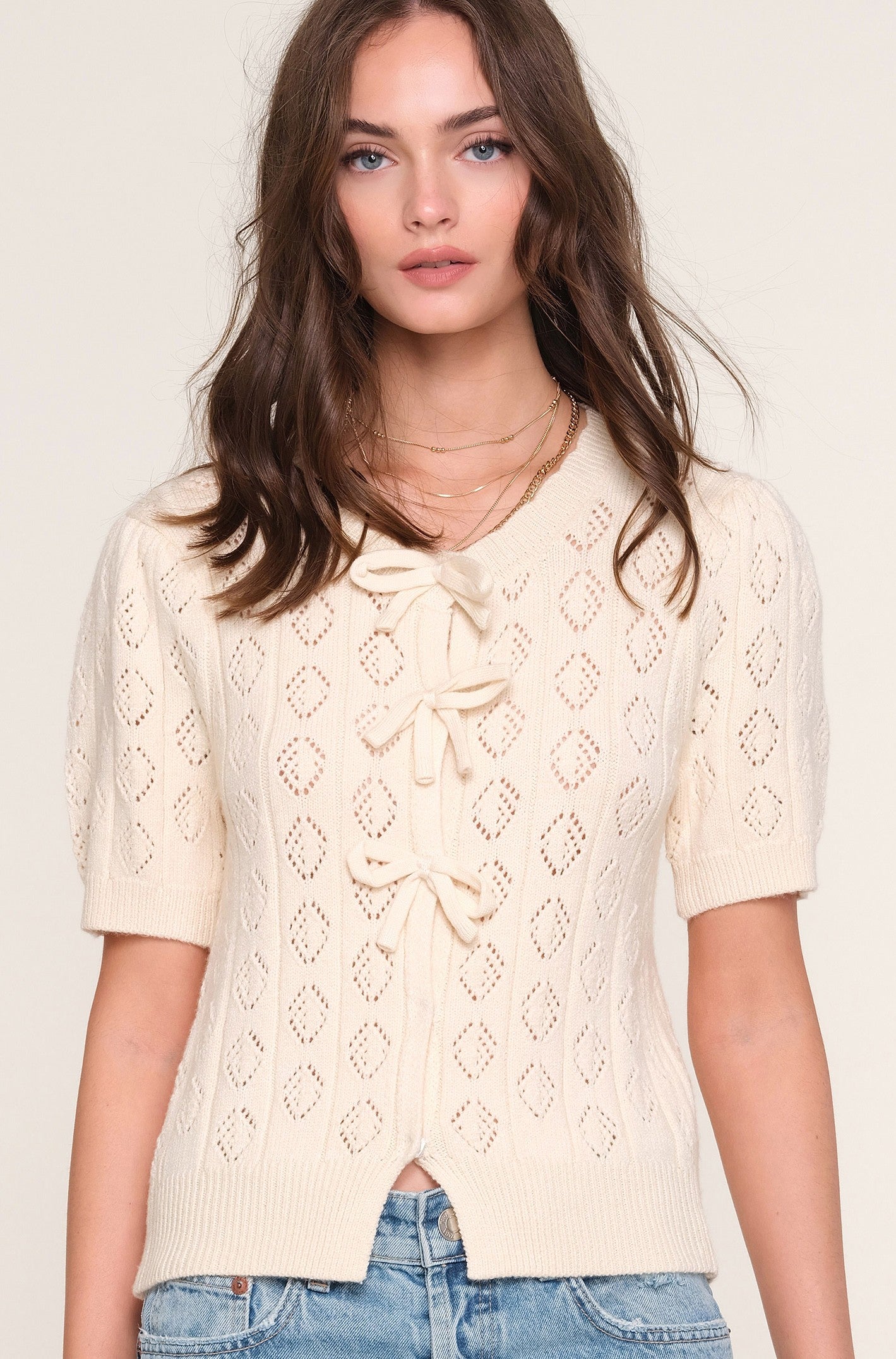 A woman wears the Heartloom Mae Cardi, a cream slim-fit knit sweater with a diamond pattern and three front bows, paired with light blue jeans against a plain background.