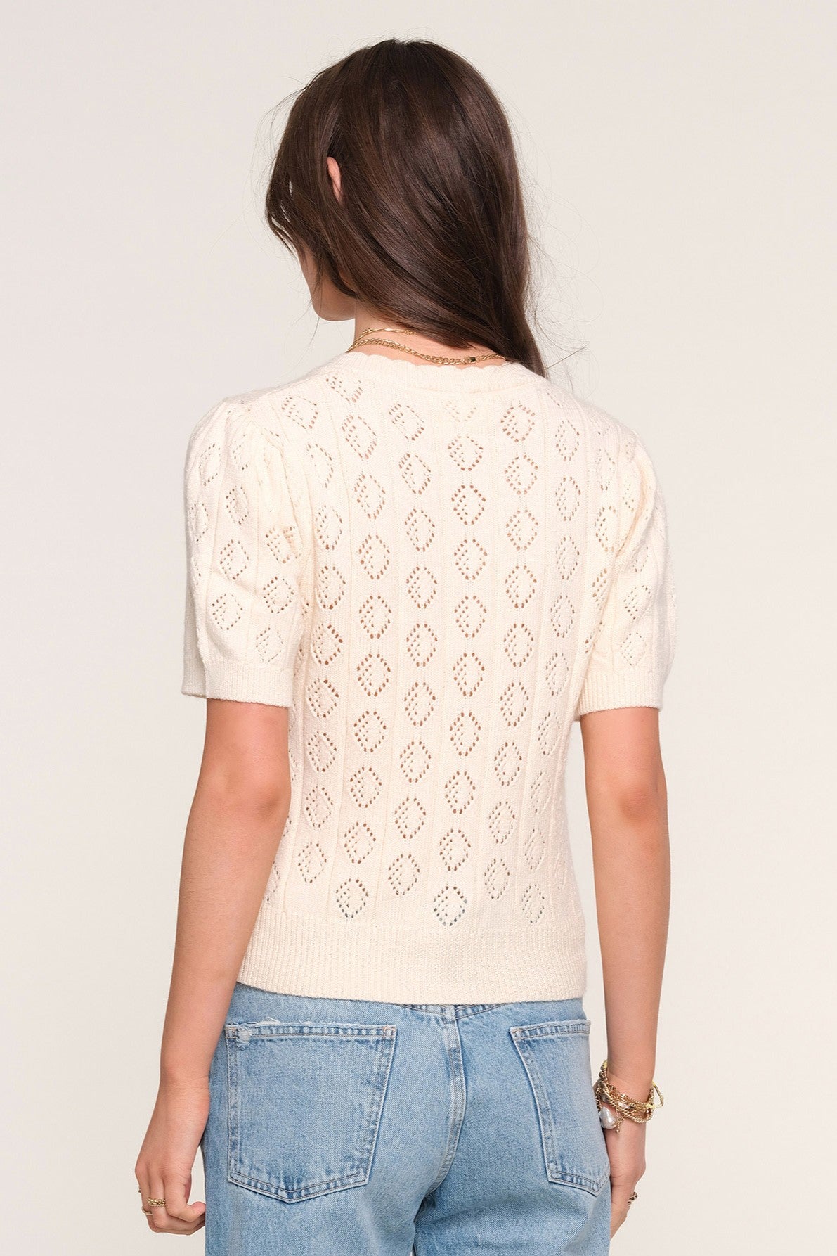 A woman is shown from the back wearing the Heartloom Mae Cardi, a cream pointelle stitch knit sweater with diamond eyelets, paired with slim light blue jeans. Her brown hair is loose and she accessorizes with bracelets and a necklace.