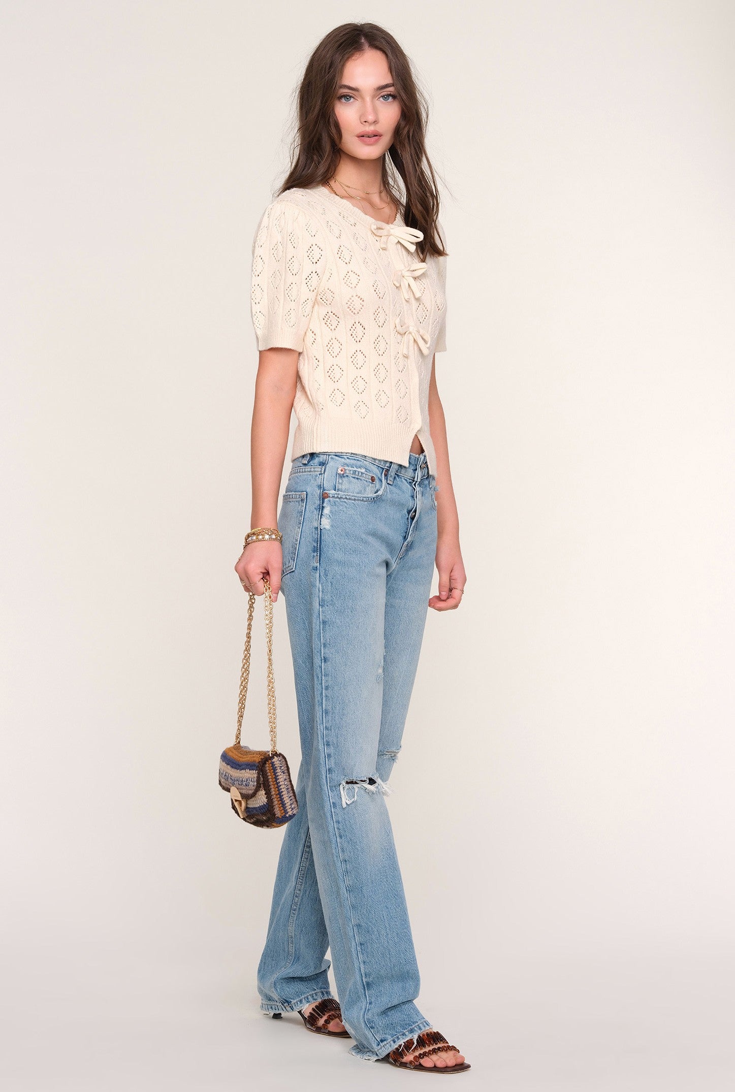 A woman stands against a plain background wearing the Mae Cardi by Heartloom, paired with light blue ripped jeans, animal print sandals, and holding a small gold-chain handbag.