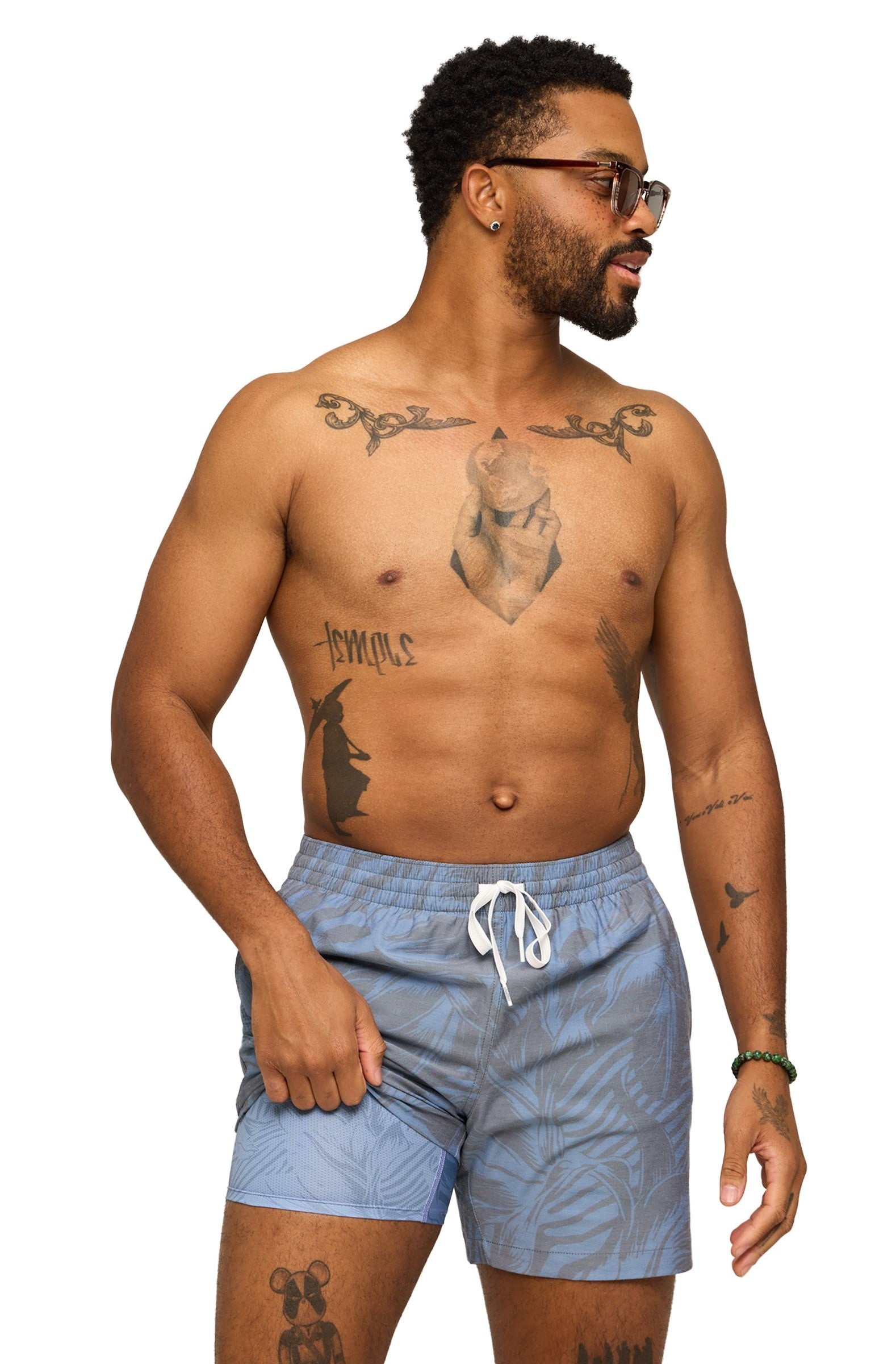 A man with short curly hair, a beard, and tattoos on his chest and arms poses shirtless in Chubbies The Indigo Crazies 7 In Lined Swim shorts, featuring a subtle leaf pattern and zippered pocket. He smiles while looking to his right.
