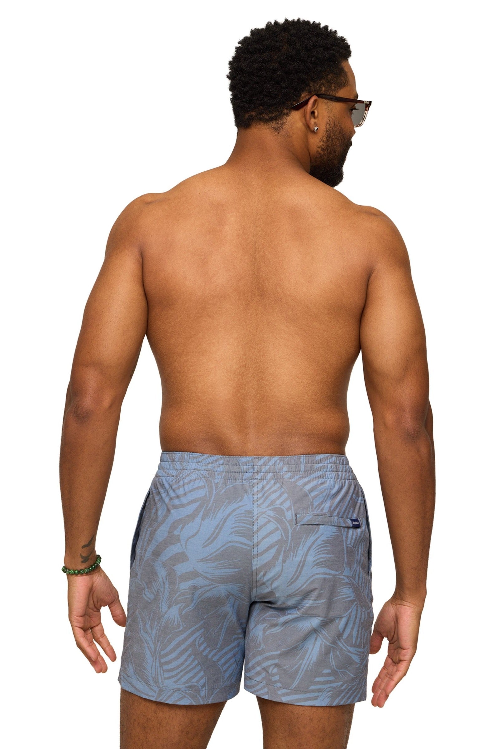 A man with short curly hair, wearing sunglasses and The Indigo Crazies 7 In Lined Swim shorts by Chubbies, stands shirtless facing away from the camera against a white background, with a bracelet visible on his left wrist.