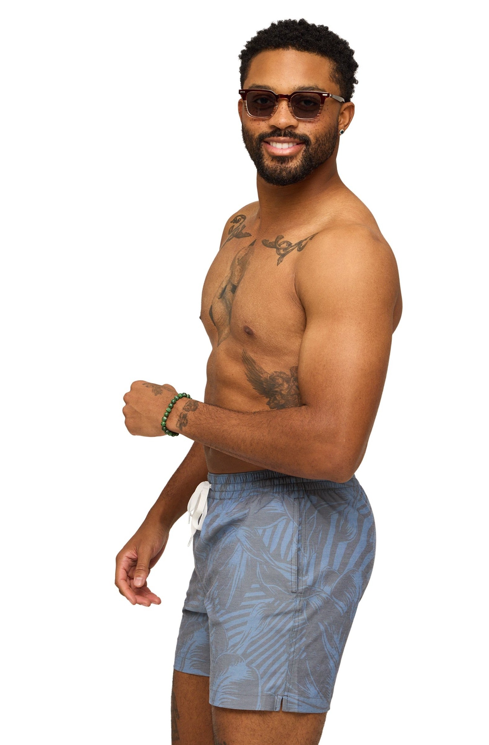 A smiling bearded man with sunglasses poses shirtless, showing tattoos, wearing Chubbies' The Indigo Crazies 7 In Lined Swim—blue patterned quick-dry shorts with four-way stretch and a zippered pocket—against a white background.