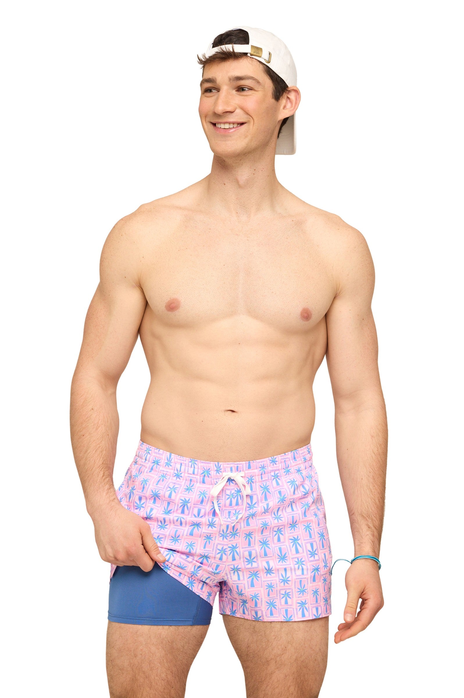 A smiling man in a backwards white cap stands shirtless, wearing Chubbies' The Miami Mosaic 5.5 Inch swim trunks with pink patterns and a visible blue compression liner. These quick-dry, four-way stretch shorts offer comfort and style on a plain white background.
