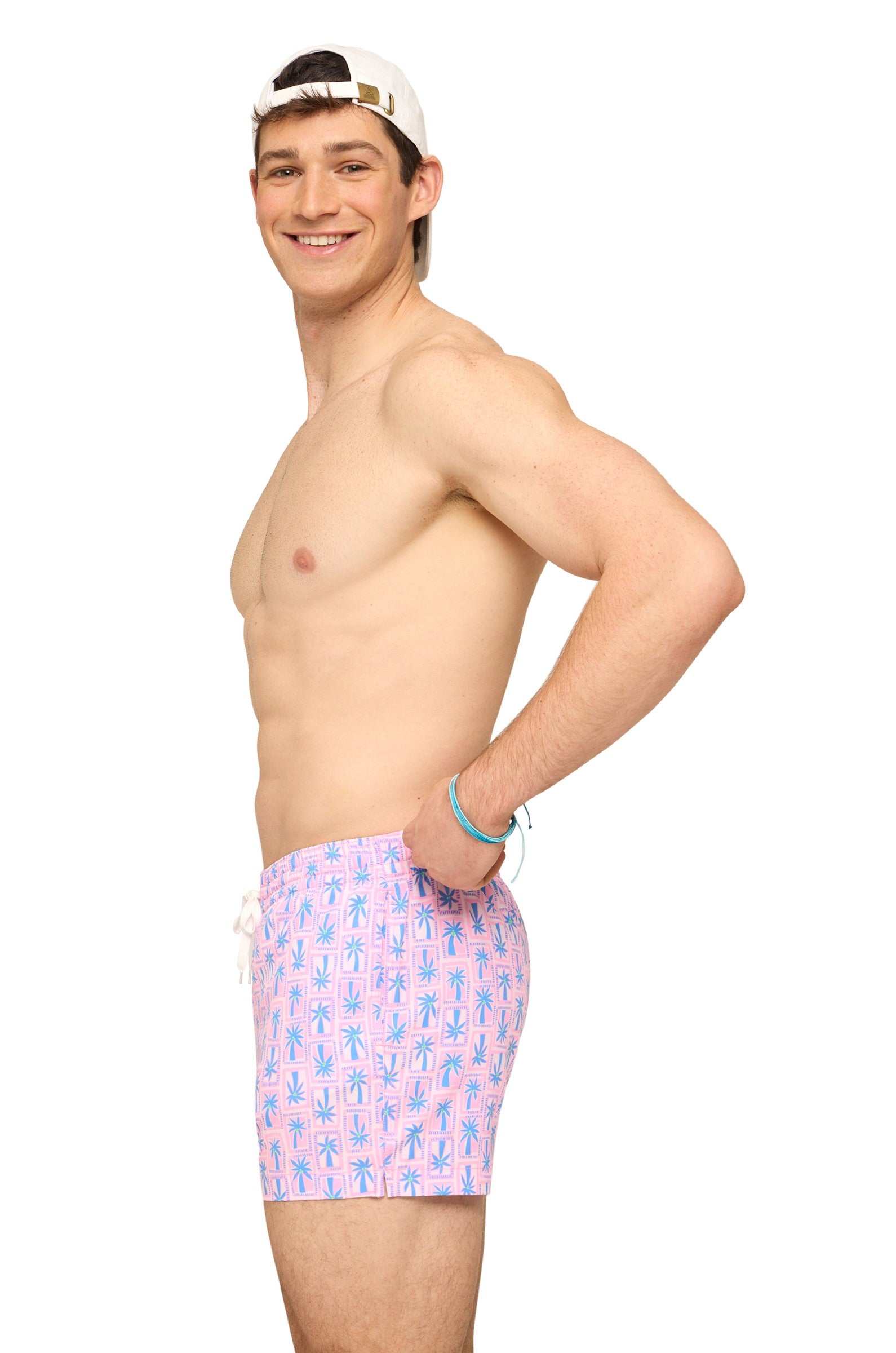 A young man in profile smiles with one hand on his hip, wearing a white backward cap and Chubbies' The Miami Mosaic 5.5 Inch quick-dry shorts with a blue palm tree pattern, set against a plain white background.