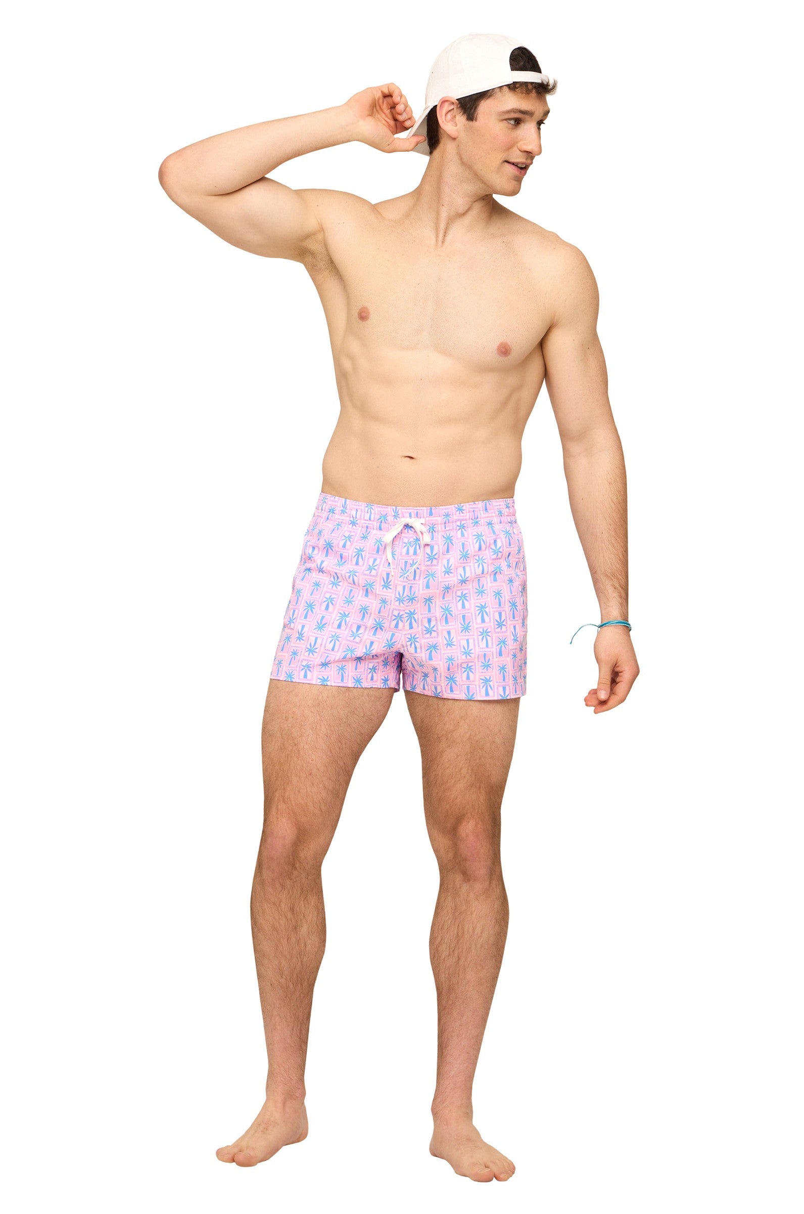 A man wearing a backward white cap and Chubbies’ The Miami Mosaic 5.5 Inch quick-dry shorts in pink with blue palm tree print smiles barefoot, with one hand behind his head, looking to his left against a white background.