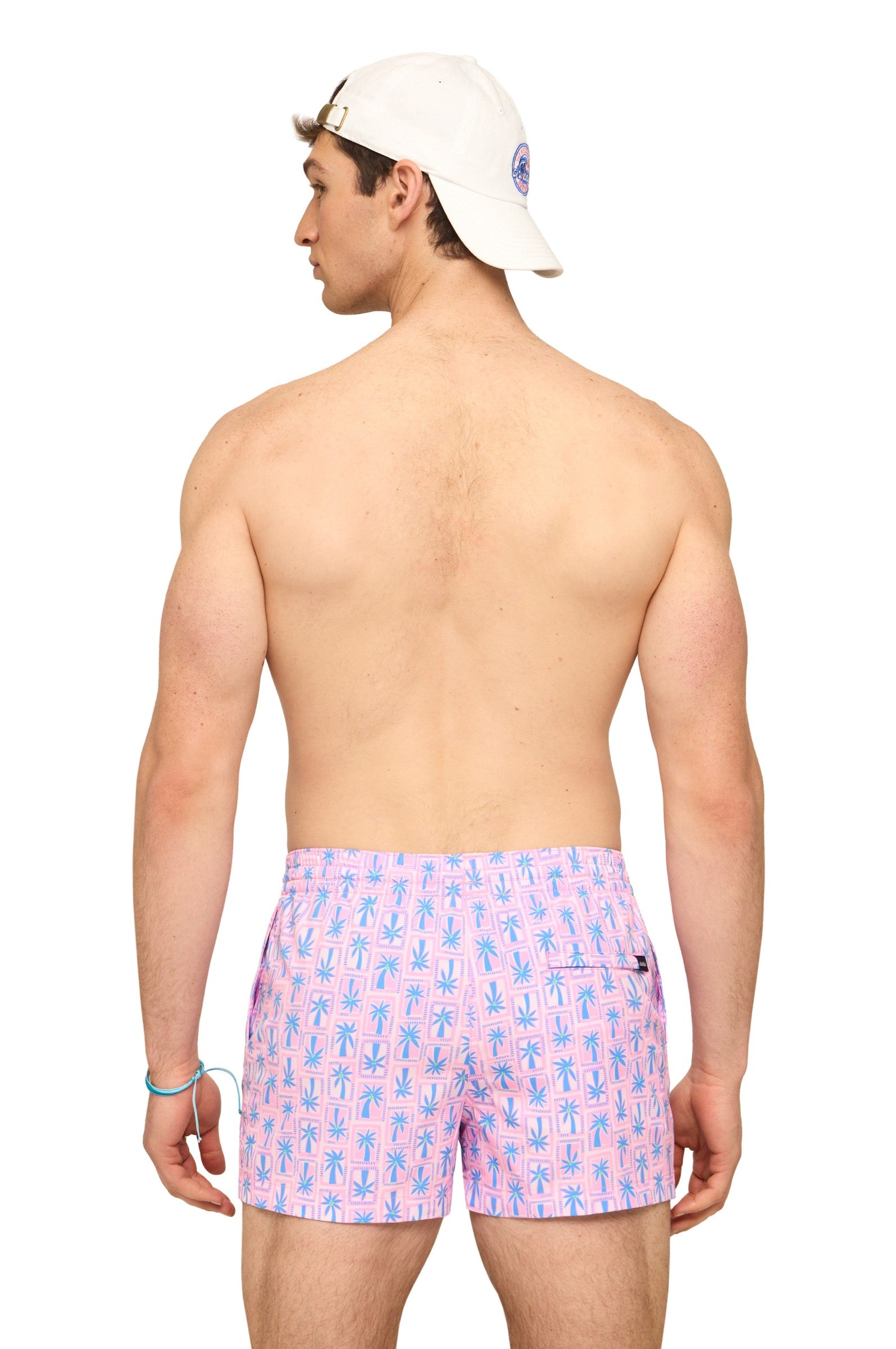 A shirtless man in a white baseball cap and Chubbies The Miami Mosaic 5.5 Inch shorts with a blue palm tree design stands with his back to the camera, showing off his shoulders.