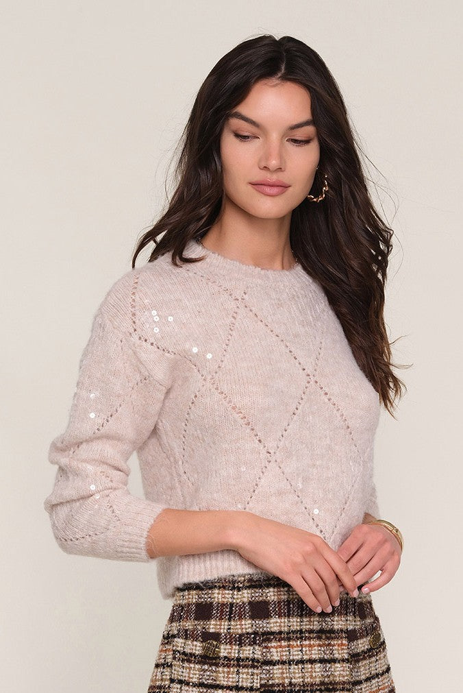 A woman with long dark hair models the Heartloom Dorian Sweater, a light beige, relaxed fit top with diamond patterns and subtle sequins, paired with a high-waisted plaid skirt against a plain light background.