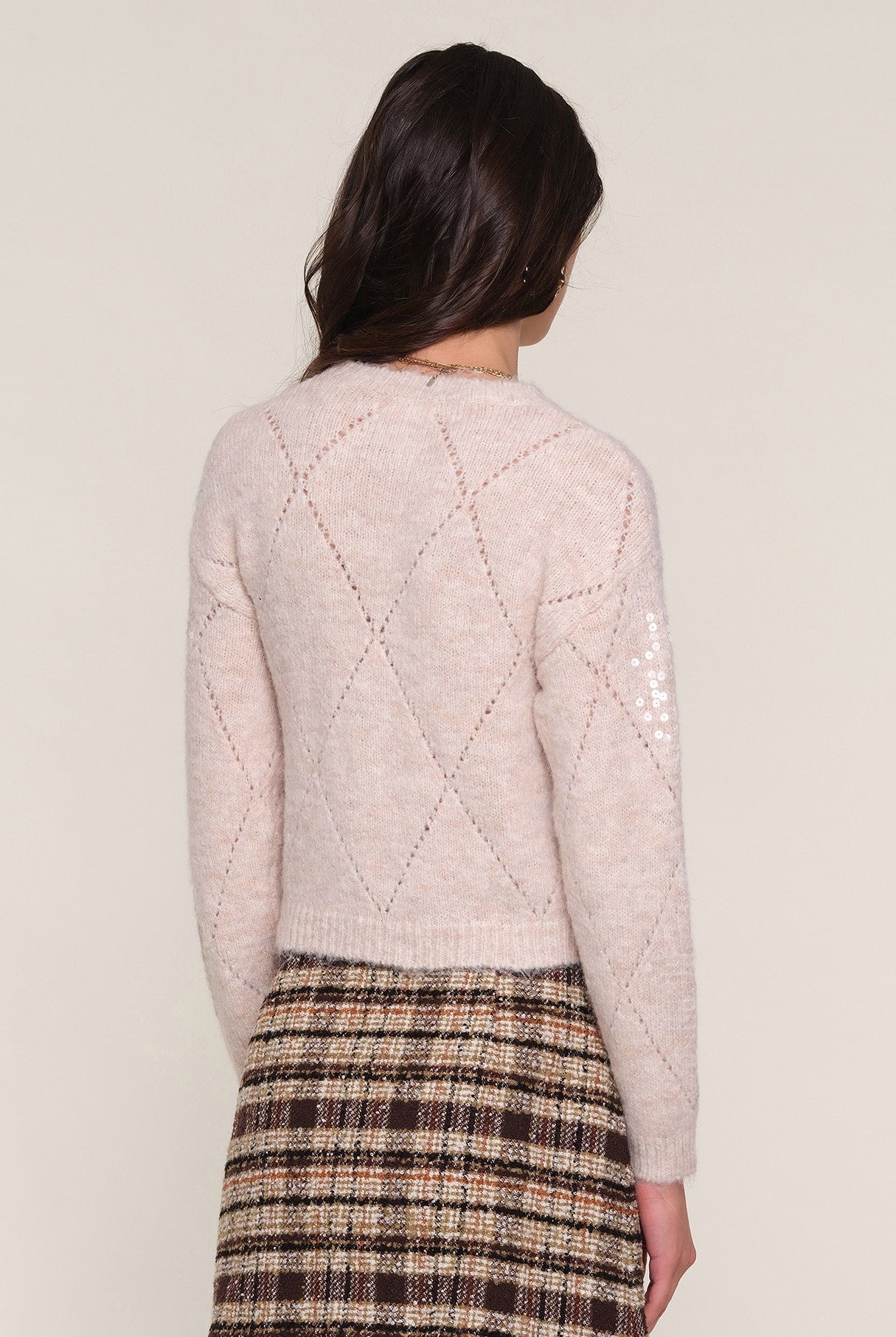 A woman with long brown hair, seen from behind, wears the Heartloom Dorian Sweater in light beige with a diamond pattern and a brown plaid skirt against a plain light background.