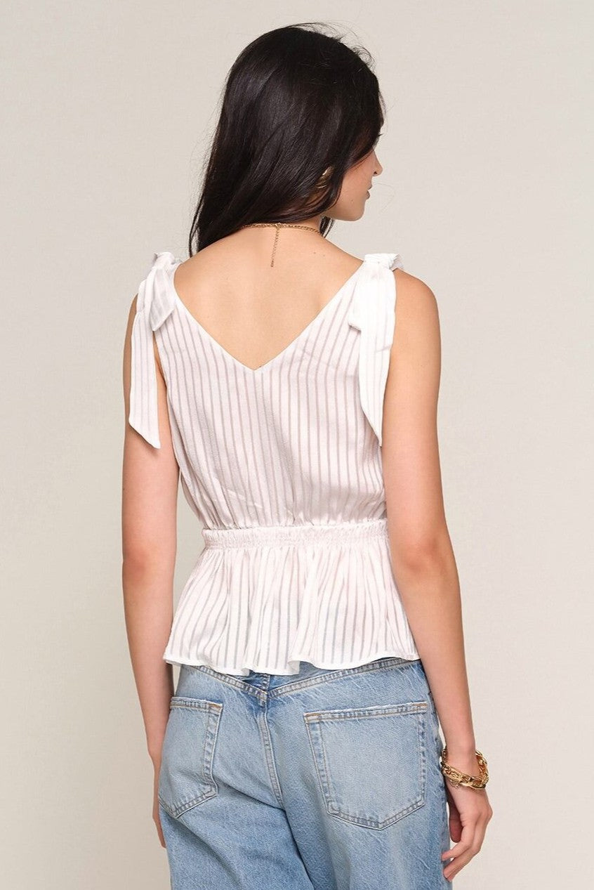 A woman with long dark hair faces away from the camera, wearing Heartloom’s Loulou Top—a slim fit, white sleeveless peplum top with vertical stripes, bow shoulder details, and a V-back—paired with light blue high-waisted jeans.