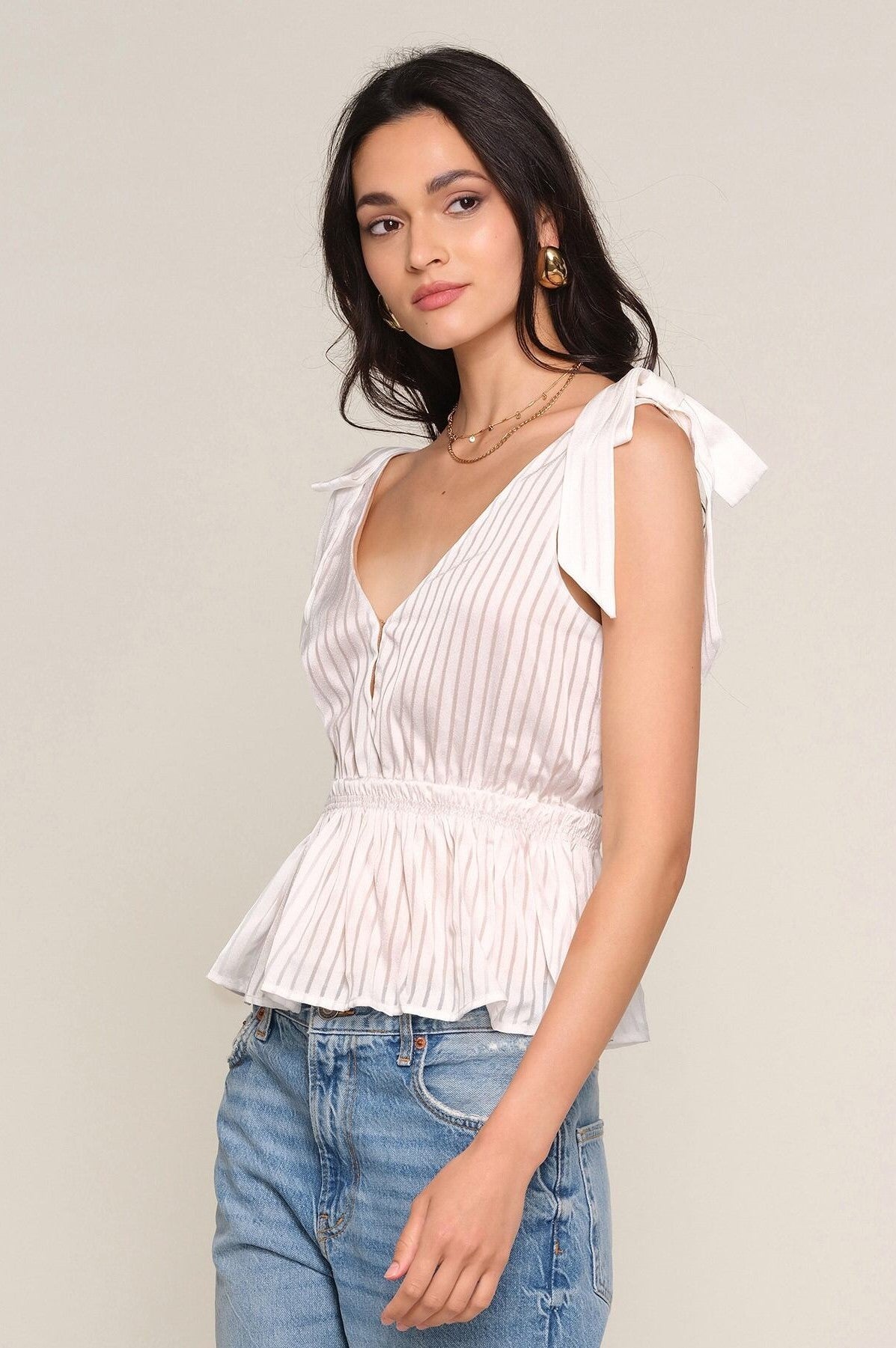 A woman wears the Heartloom Loulou Top—a sleeveless, white satin peplum with vertical stripes, V-neck, and tie straps—paired with blue jeans as she stands against a plain light background.