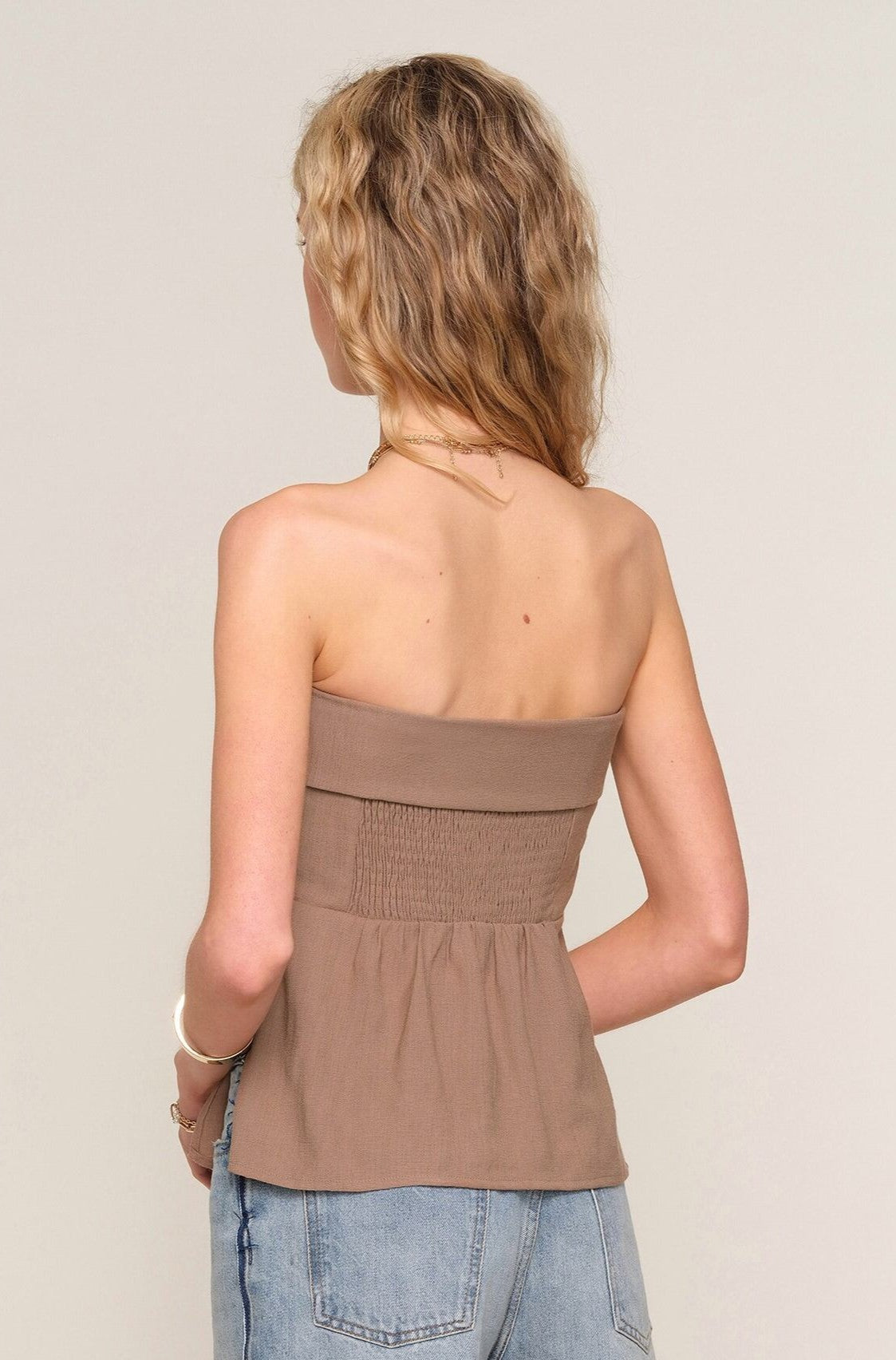 A woman with wavy blonde hair is seen from the back wearing the Heartloom Anne Top in taupe, a strapless style with a smocked waist and ruched back detail, paired with light blue jeans. She accessorizes with bracelets and a necklace.