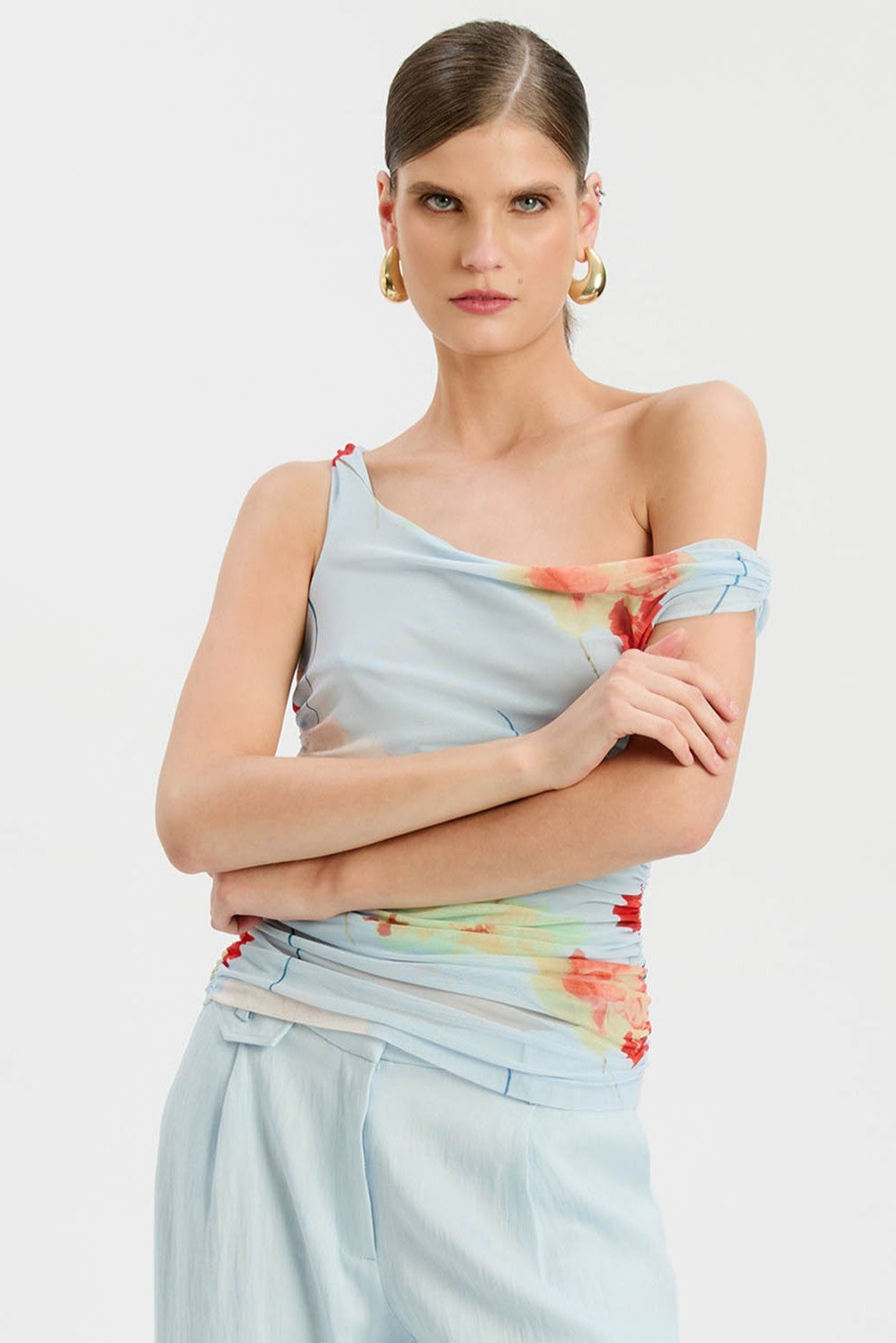 A woman with slicked-back hair wears Bardot's Layana Printed Mesh Asym Top in baby blue floral, paired with matching blue pants and gold hoop earrings, standing arms crossed against a plain white background.