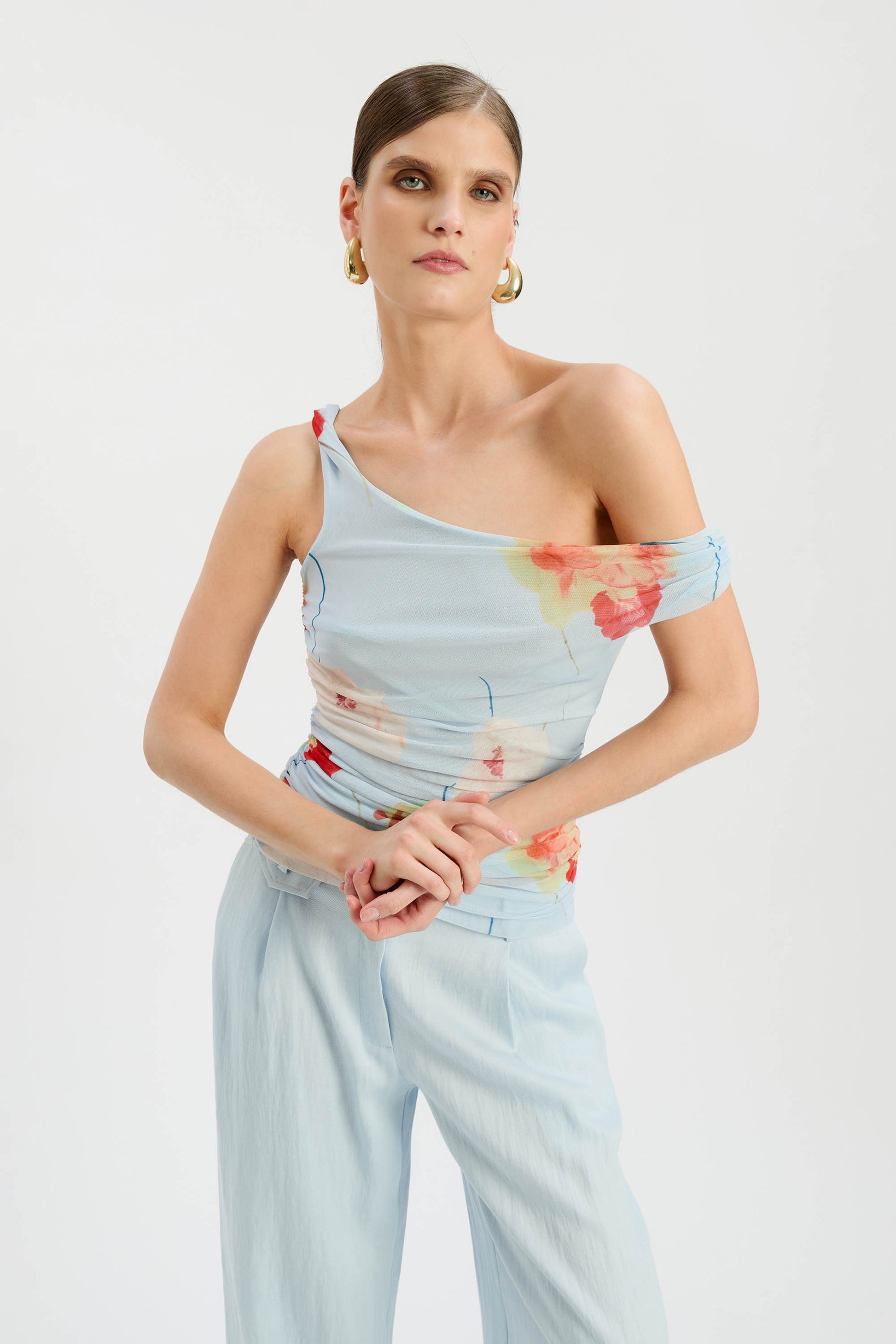 A woman models the Bardot Layana Printed Mesh Asym Top, featuring a baby blue floral design and asymmetric neckline, paired with light blue pants. She completes her semi-formal look with gold hoop earrings and slicked-back hair.