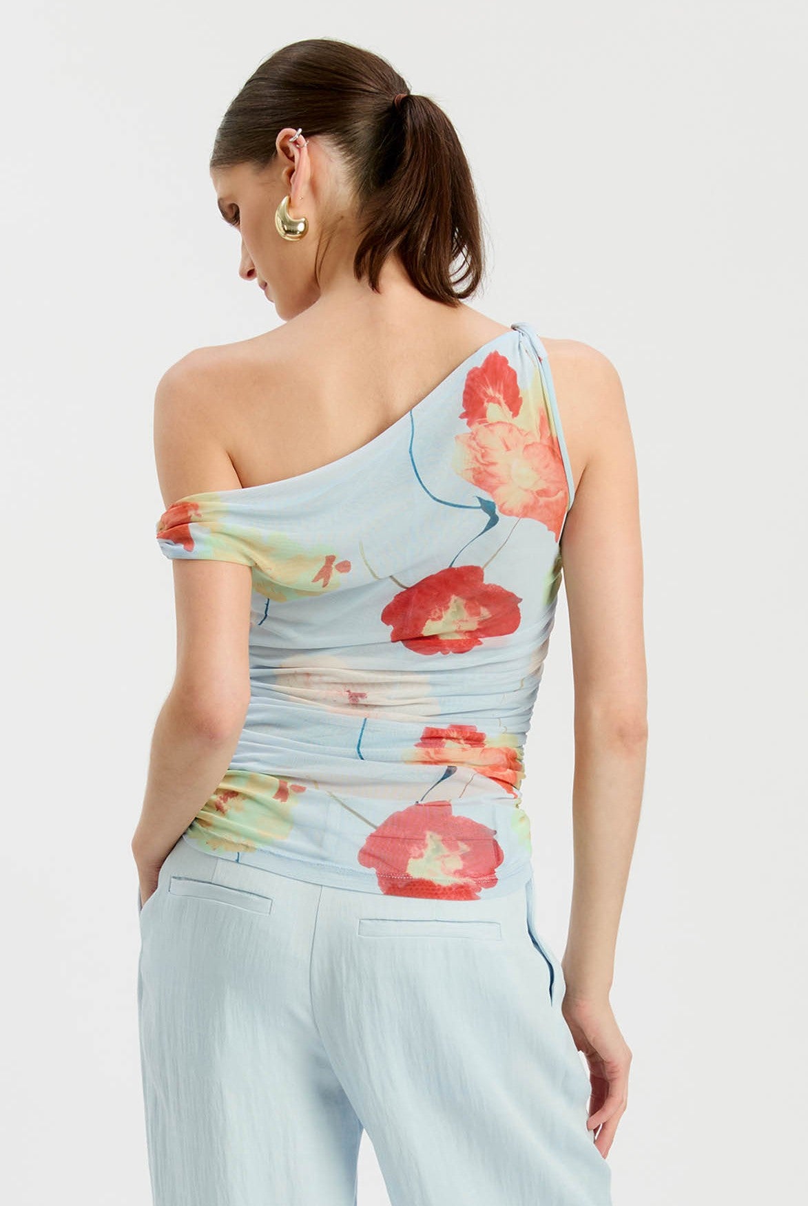 A woman seen from behind wears the Bardot Layana Printed Mesh Asym Top in baby blue, paired with light blue trousers.