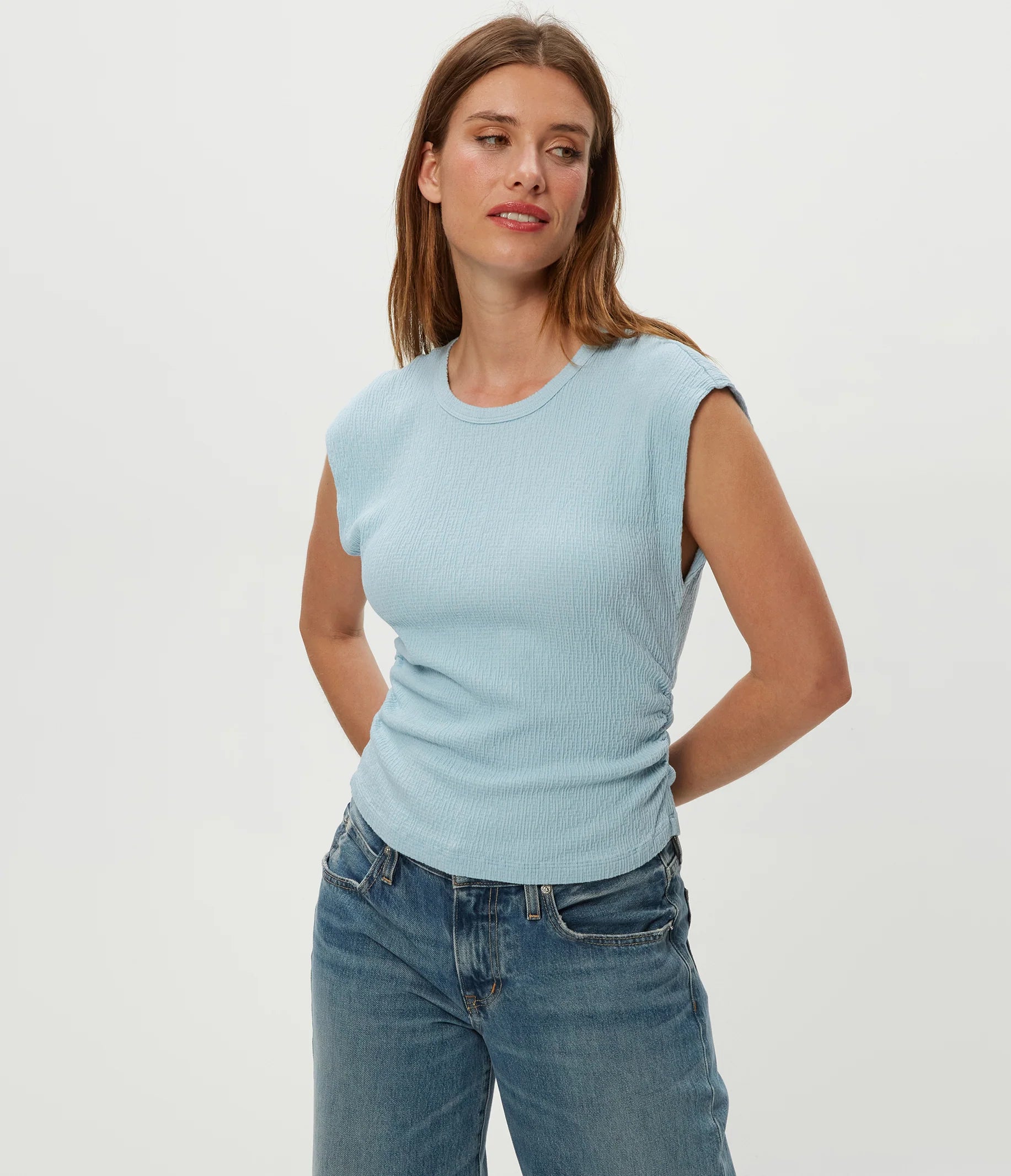 A woman with straight brown hair wears the Michael Stars Leighton Power Shoulder Top in Fountain and blue jeans. She stands against a white background, hands behind her back, looking to the side and smiling softly.