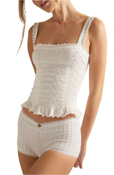 A woman wears the Free People So Soft Femme Square Top, a white knit tank with ruffled edges, paired with matching high-waisted shorts featuring a textured pattern and a small bow at the waistband.