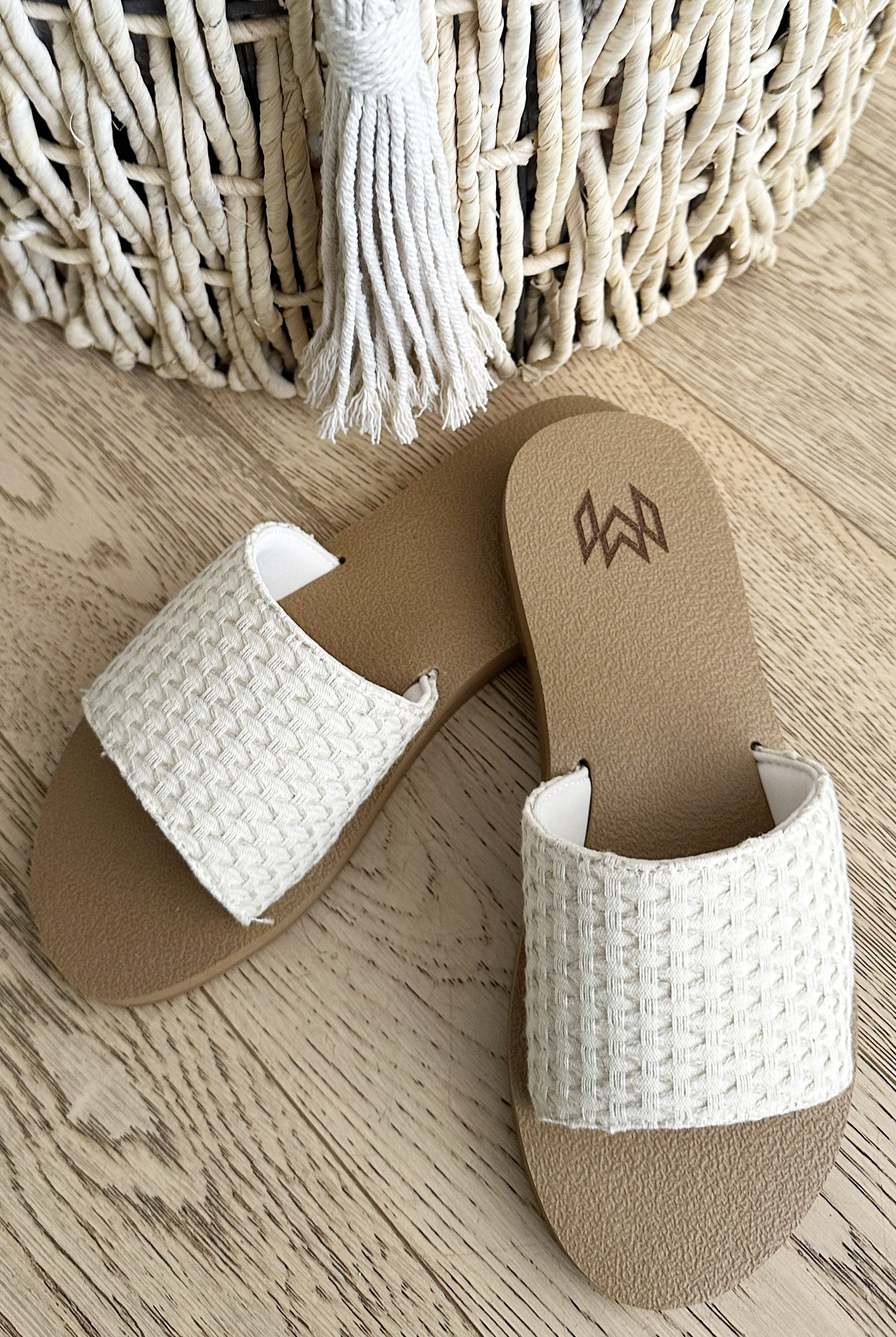 The Malvados Ellie 2- Hampton beige slide sandals with woven white straps and a memory foam footbed are placed on a light wooden floor next to a woven basket with white tassels.