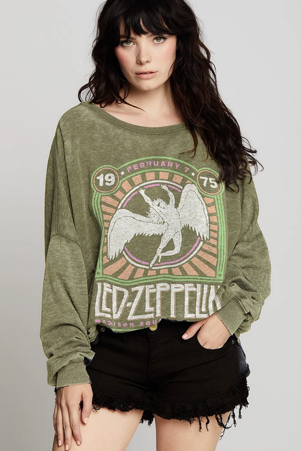 A woman with long dark hair stands facing forward against a plain background, wearing the Recycled Karma Led Zeppelin Msg Ls olive green sweatshirt with an oversized, vintage-inspired drape and black frayed shorts.